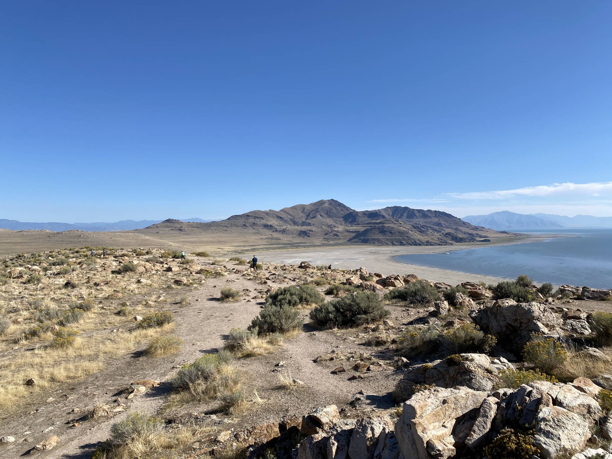 A real photo from Buffalo Point Trail on Antelope Island showing a rocky hillside trail with the Great Salt Lake and distant mountains under warm evening light