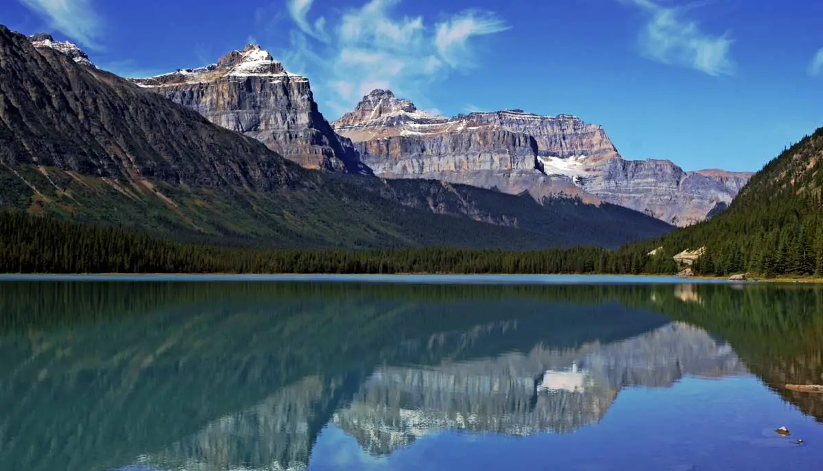 A real photo from a roadside viewpoint overlooking Waterfowl Lakes with deep blue water, a line of dark evergreens, and a distant glacier-capped mountain range