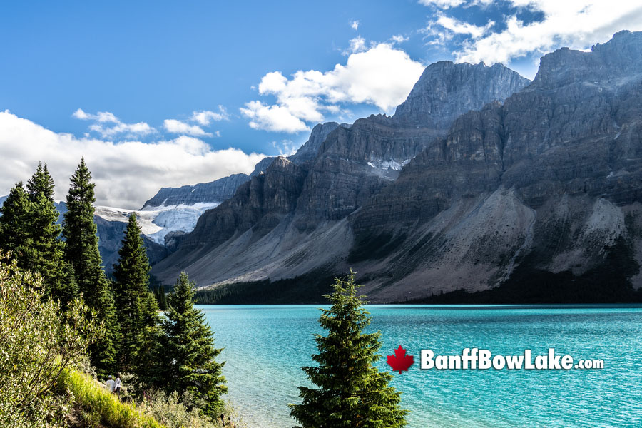 A real photo of Bow Lake in Banff National Park with turquoise water, a rocky shoreline in the foreground, and a steep mountain rising behind the lake under scattered clouds