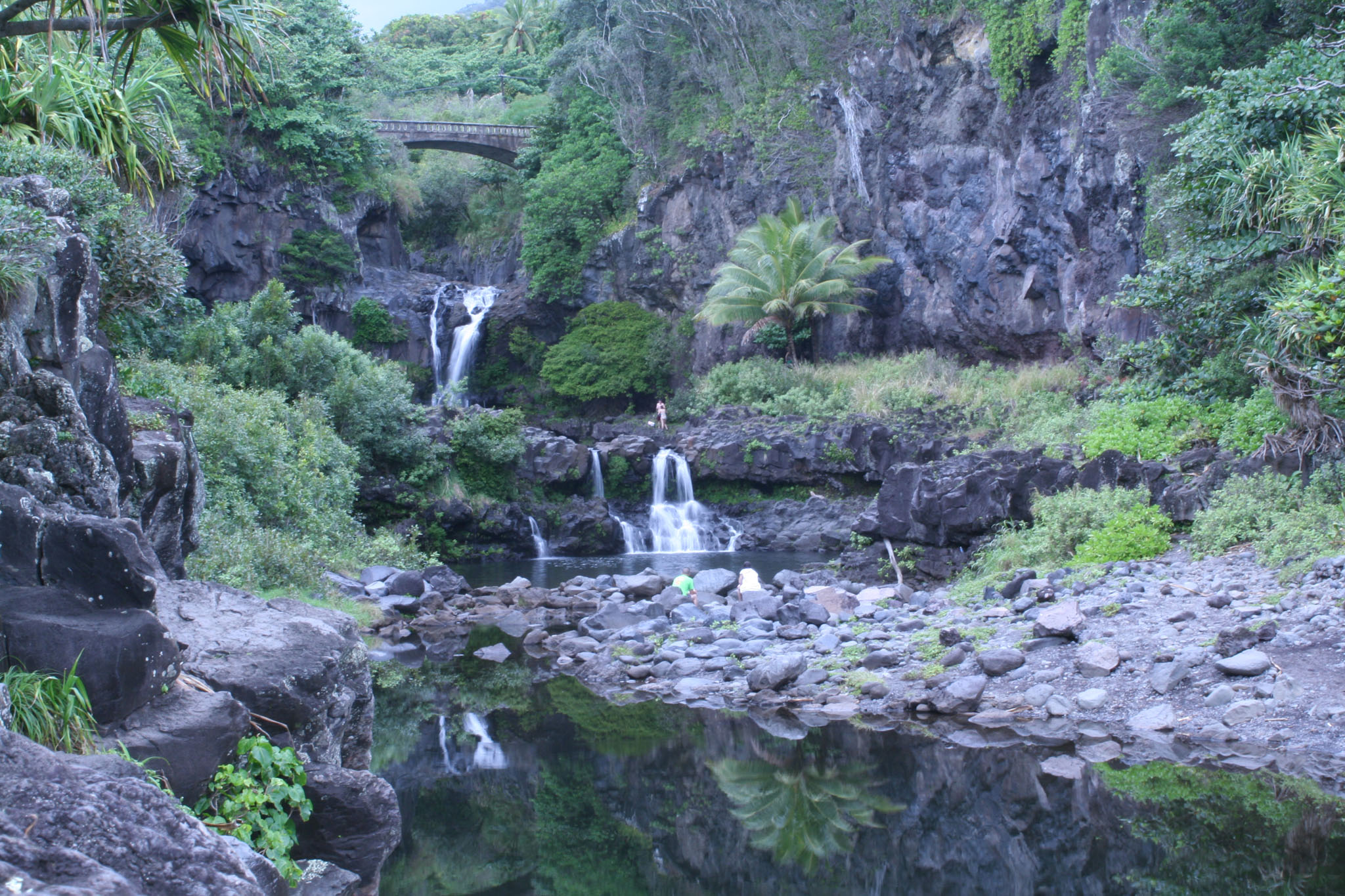 A real photo of ʻOheʻo Gulch in Kīpahulu, Maui with cascading water over dark rock steps and green vegetation along the stream, no text
