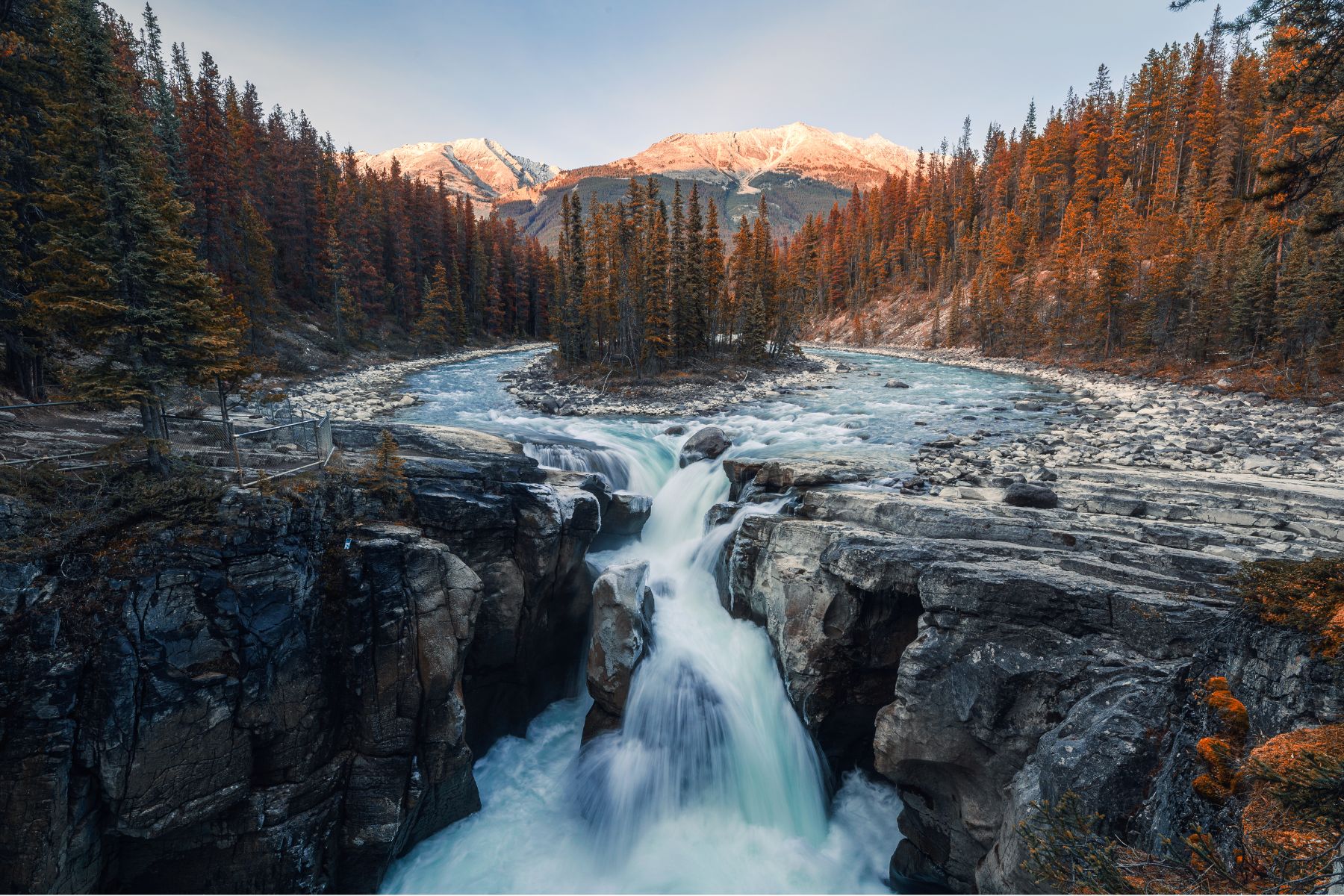 A real photo of Sunwapta Falls with white water rushing through a rocky canyon, evergreen trees framing the scene, and a cloudy mountain sky overhead