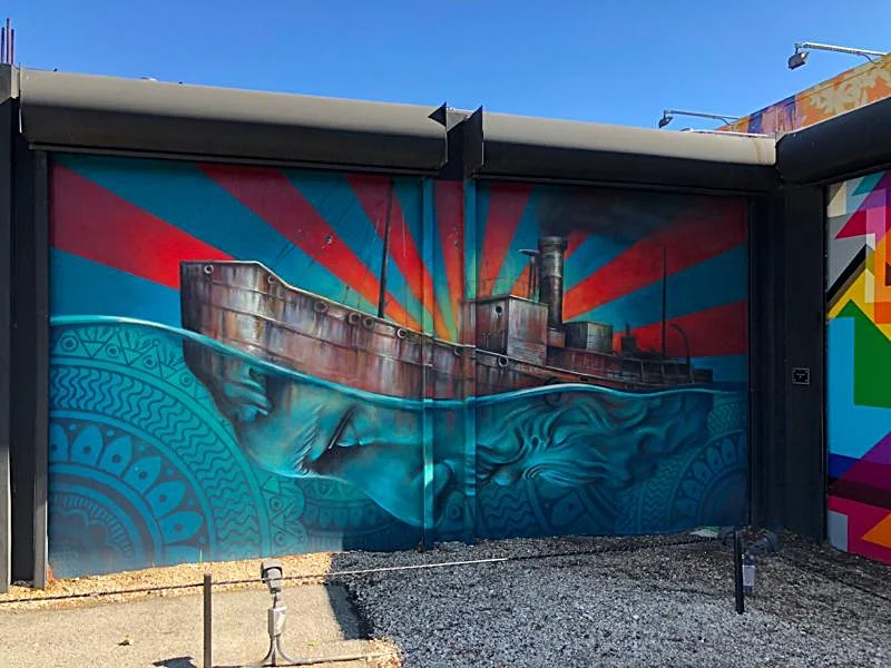 A real photo of a large colorful street art mural painted on a Wynwood warehouse wall at dusk with a few people walking by