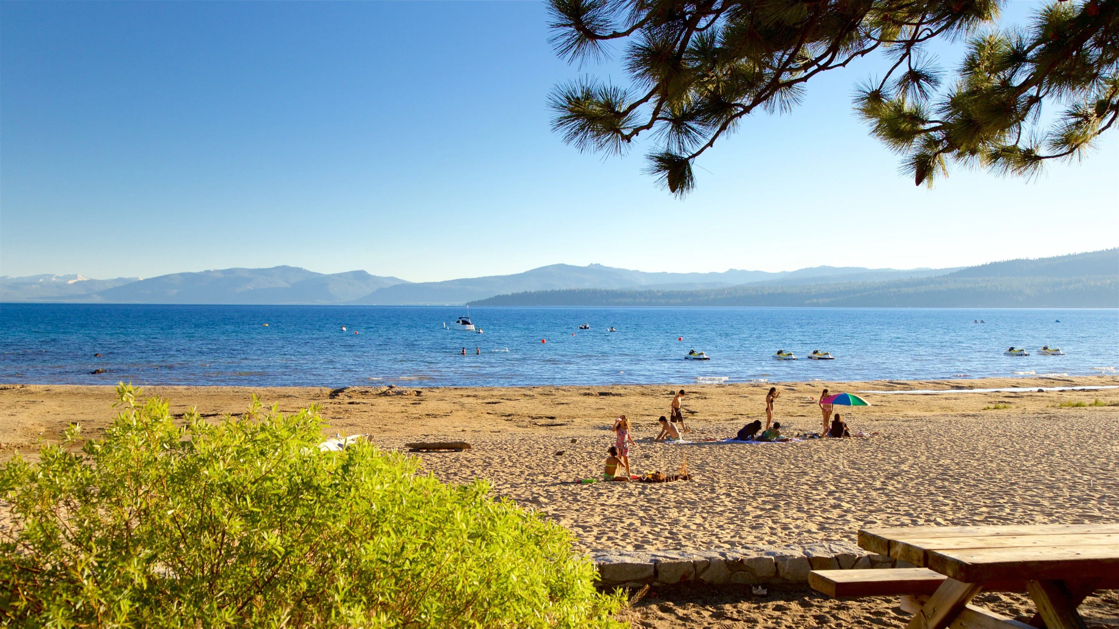 A real photo of a sandy shoreline walk at Kings Beach with people strolling near the water and forested slopes in the background