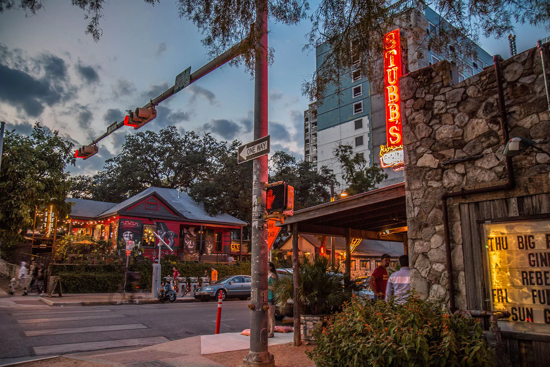 A real photo of an Austin music venue exterior in the Red River Cultural District at dusk, with a line of people waiting under marquee lights