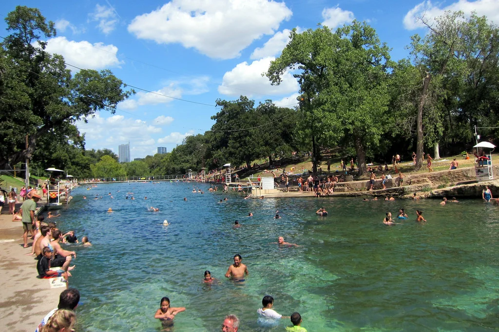 A real photo of swimmers in the clear water at Barton Springs Pool on a sunny day, with limestone edges and trees in the background