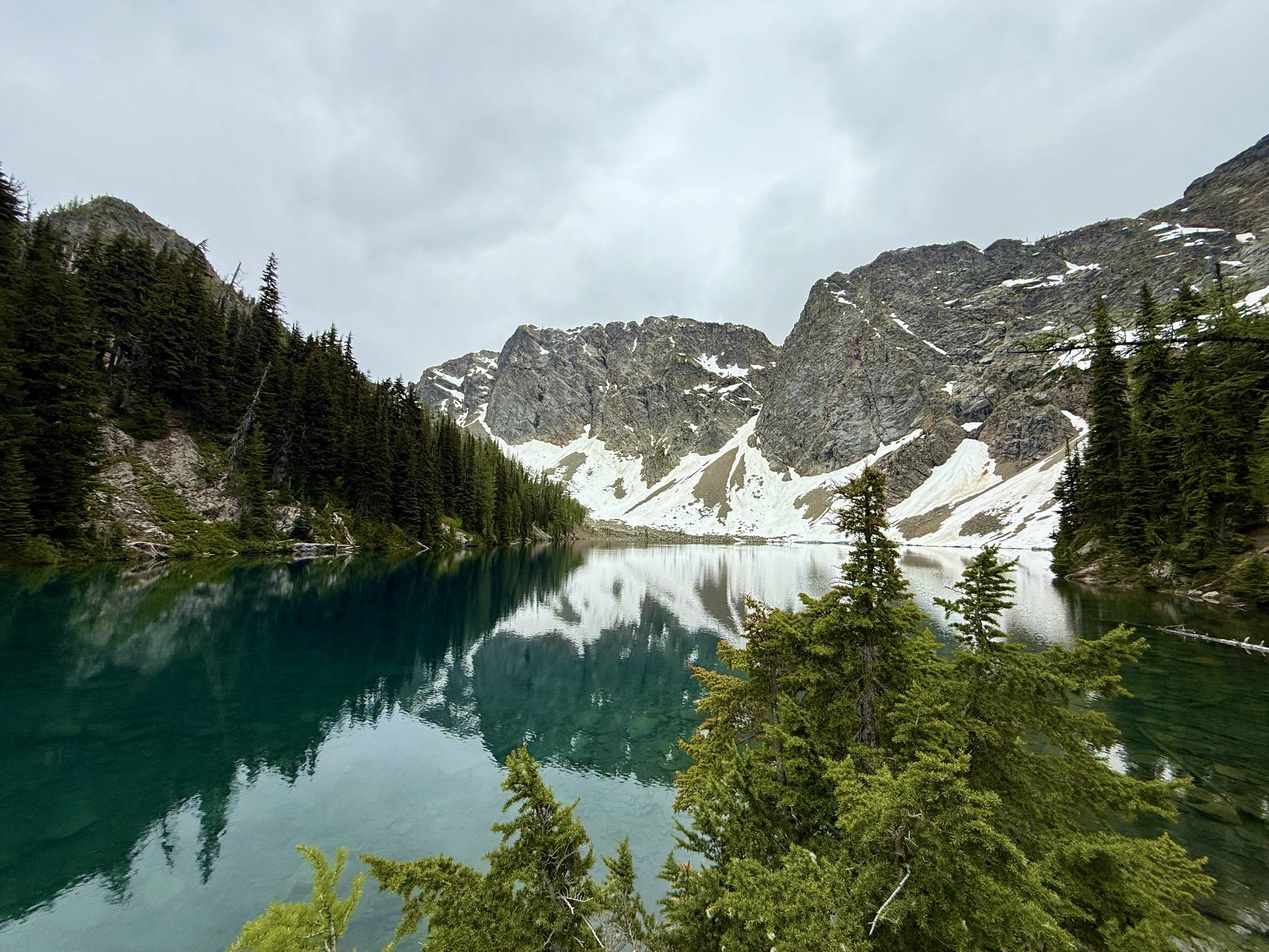 A real photograph at Blue Lake in the North Cascades showing clear blue water at the rocky shoreline, subalpine trees nearby, and steep mountain slopes in the background
