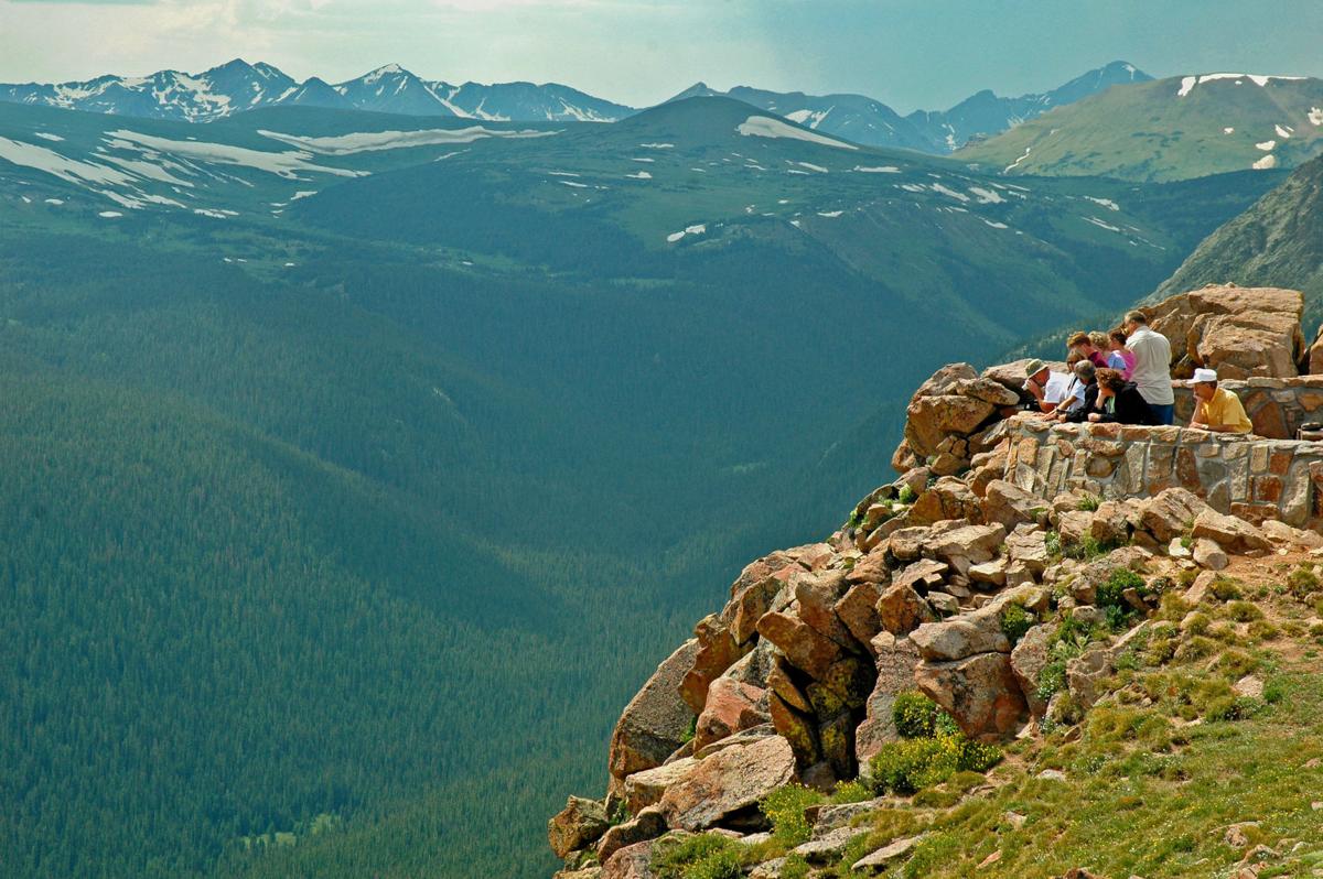 A real photograph at Forest Canyon Overlook on Trail Ridge Road, with visitors standing at a viewpoint looking across a deep canyon toward rugged alpine mountains