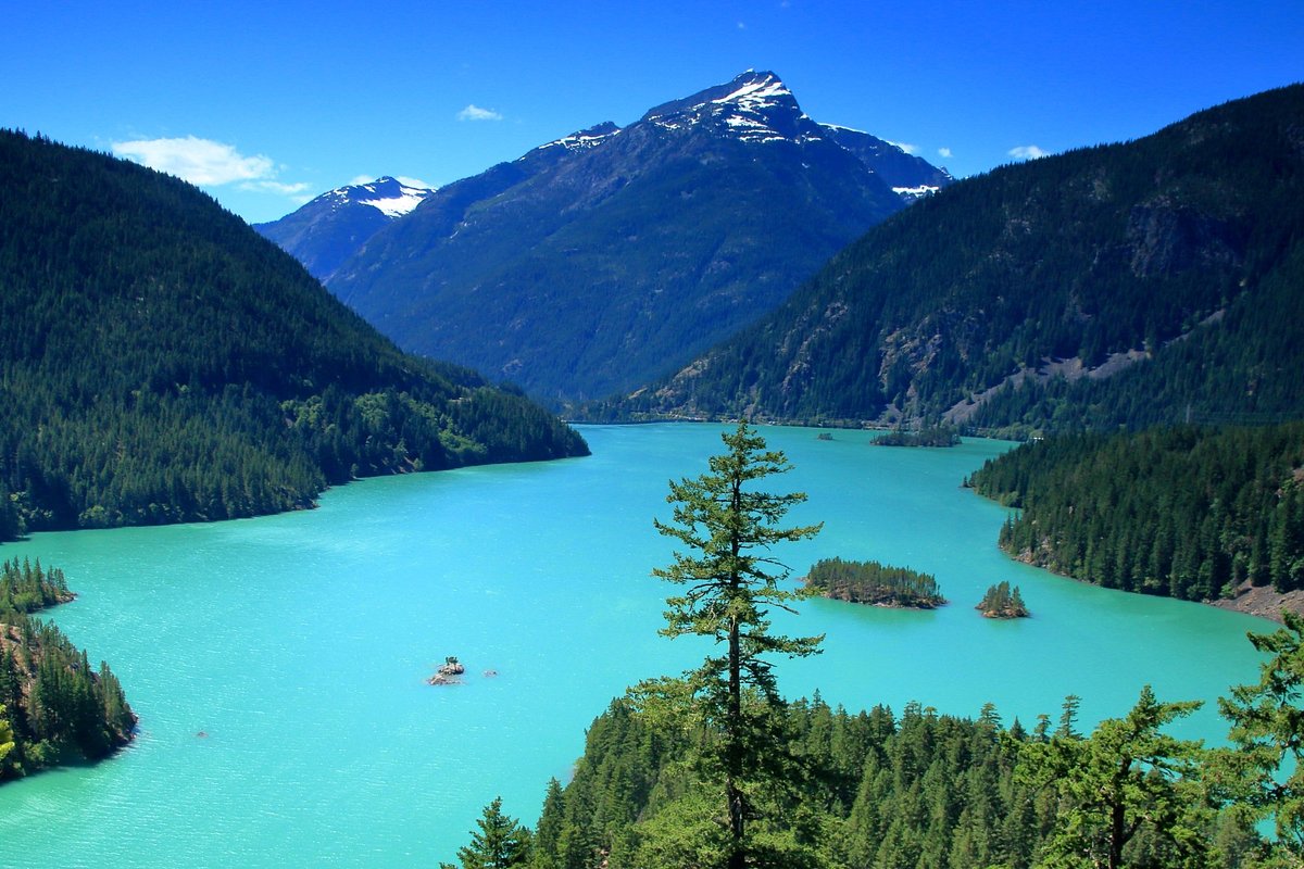 A real photograph from Diablo Lake Vista Point showing bright turquoise Diablo Lake below, surrounded by dark evergreen slopes and rugged North Cascades peaks under a partly cloudy sky