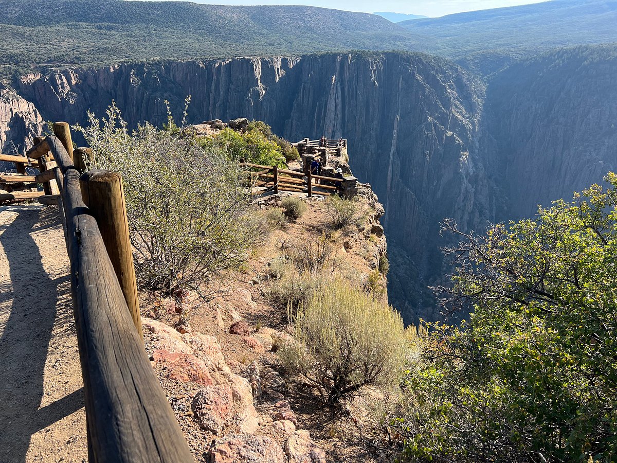 A real photograph from Gunnison Point overlook on the South Rim with a stone or metal railing in the foreground and the narrow canyon opening out across rugged mesas