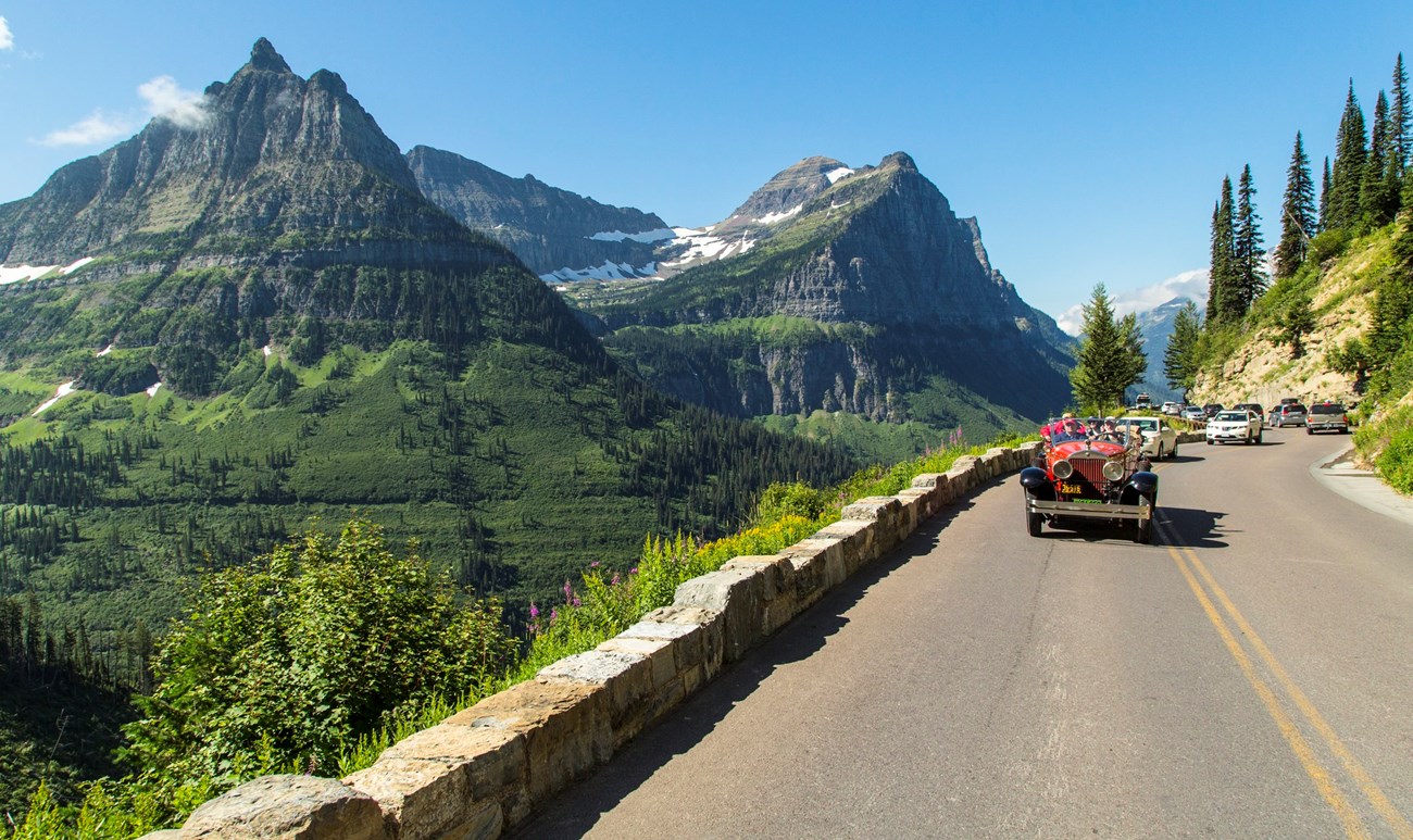 A real photograph from Logan Pass on Going-to-the-Sun Road in Glacier National Park, with rugged mountain peaks, patches of snow, and a winding road far below under a bright summer sky