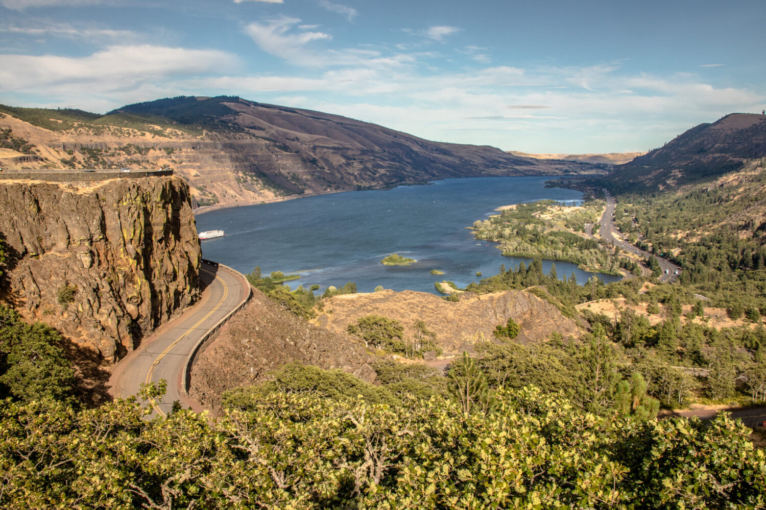 A real photograph from Rowena Crest showing the sweeping curves of the Historic Columbia River Highway below, golden hillside slopes, and the Columbia River stretching into the distance