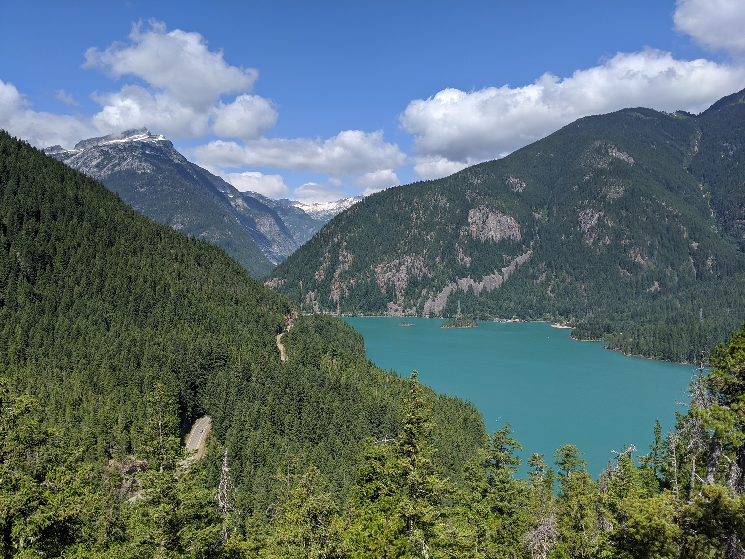 A real photograph from Thunder Knob showing a high view over Diablo Lake with evergreen forests, layered mountain ridges, and soft afternoon light