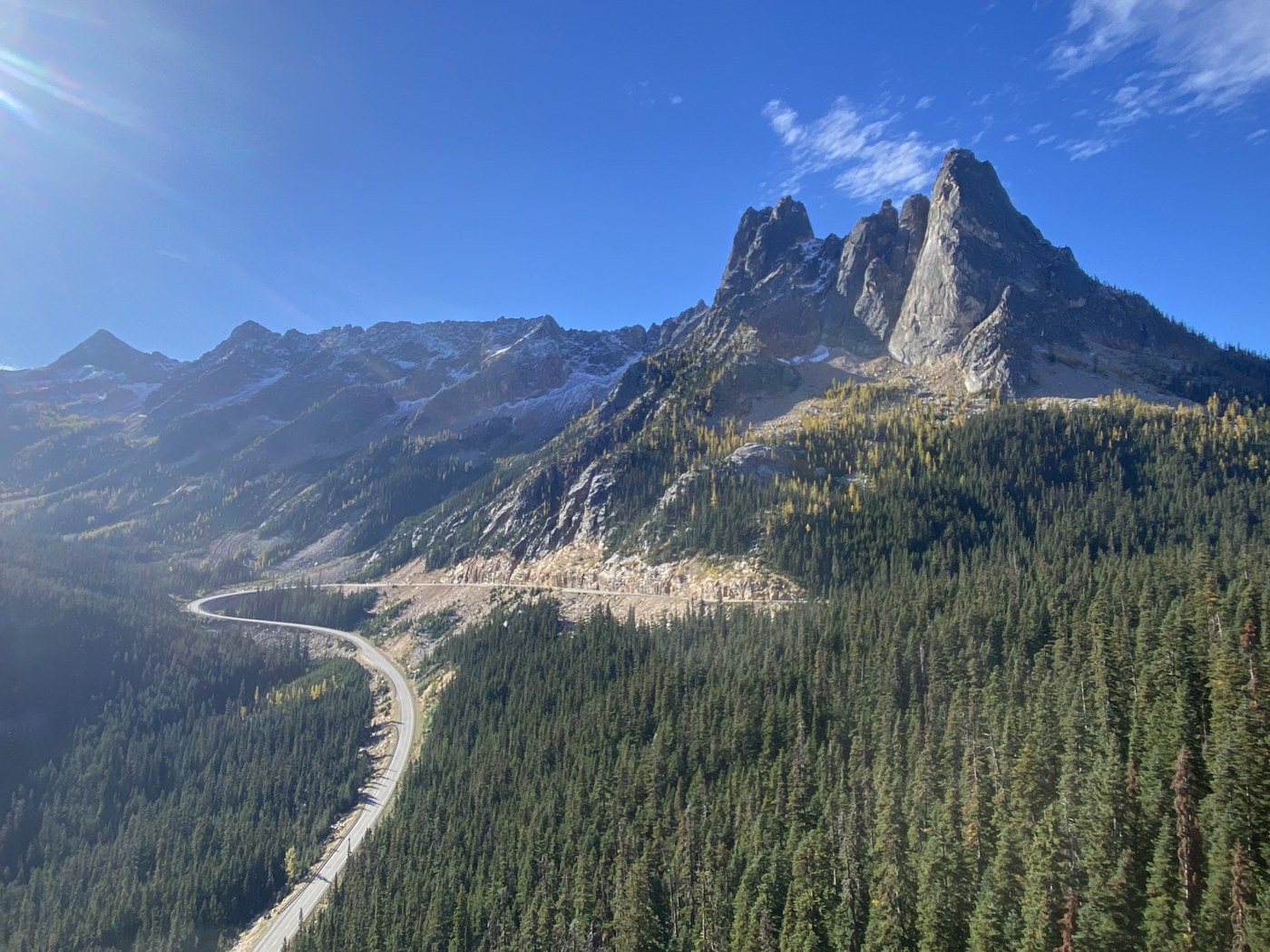 A real photograph from Washington Pass Overlook with Liberty Bell Mountain towering above, a paved path in the foreground, and evergreen trees lining the viewpoint