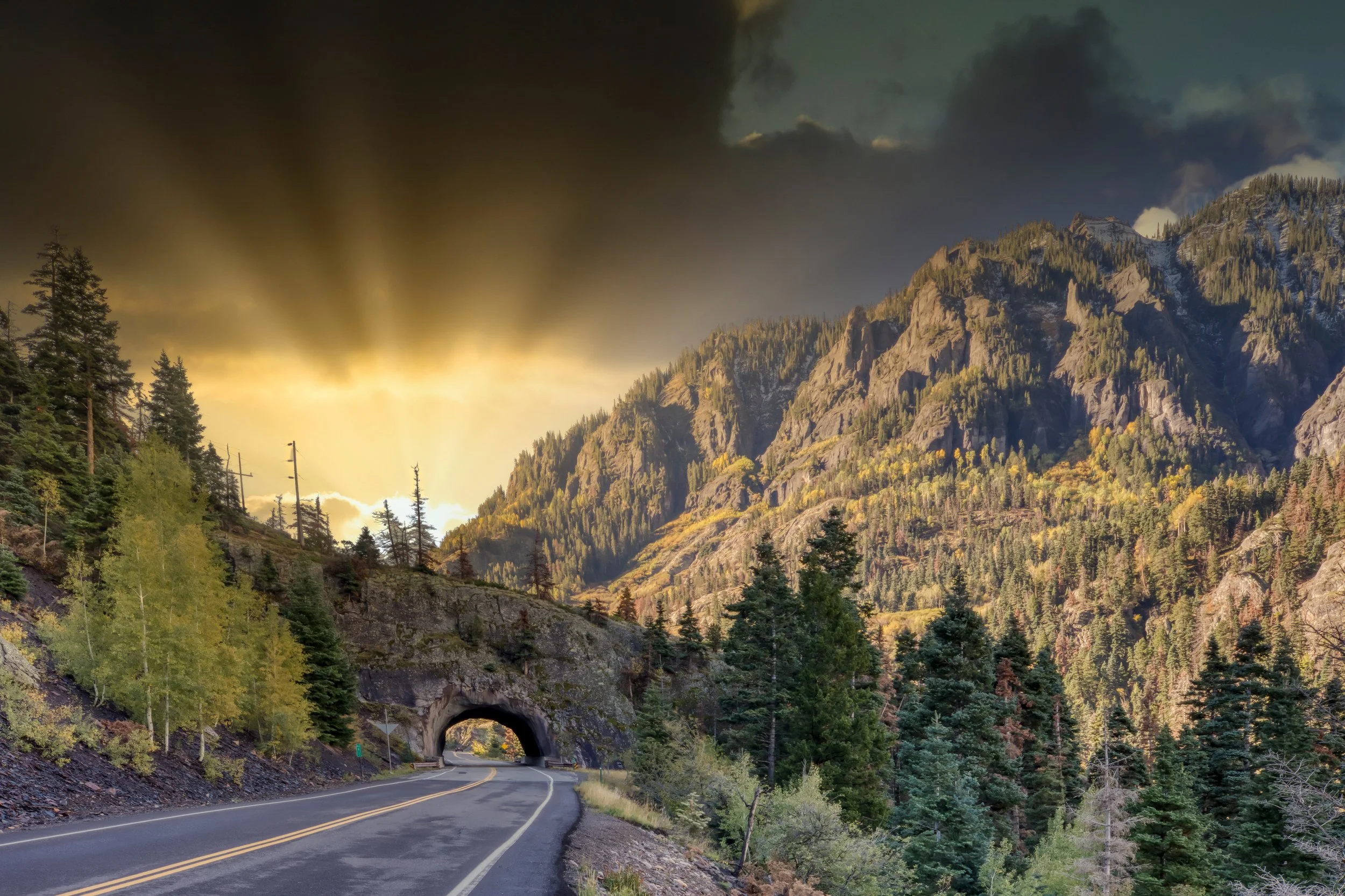 A real photograph from a roadside overlook on US 550 near Ouray showing a winding mountain highway, steep cliffs, and layered San Juan peaks under blue sky
