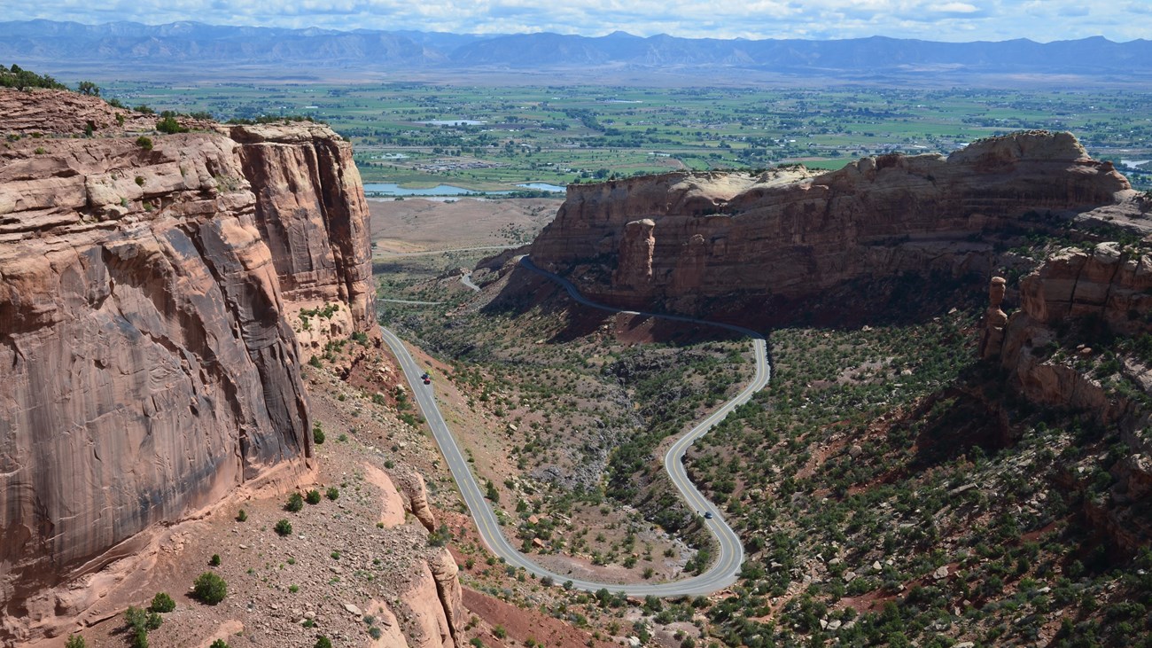 A real photograph from a roadside pullout on Rim Rock Drive showing layered red canyon walls and a winding valley far below under bright midday sun