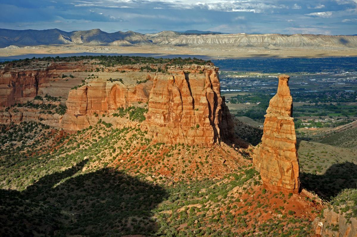 A real photograph from an overlook in Colorado National Monument showing the tall Independence Monument spire rising above red canyon walls with blue sky
