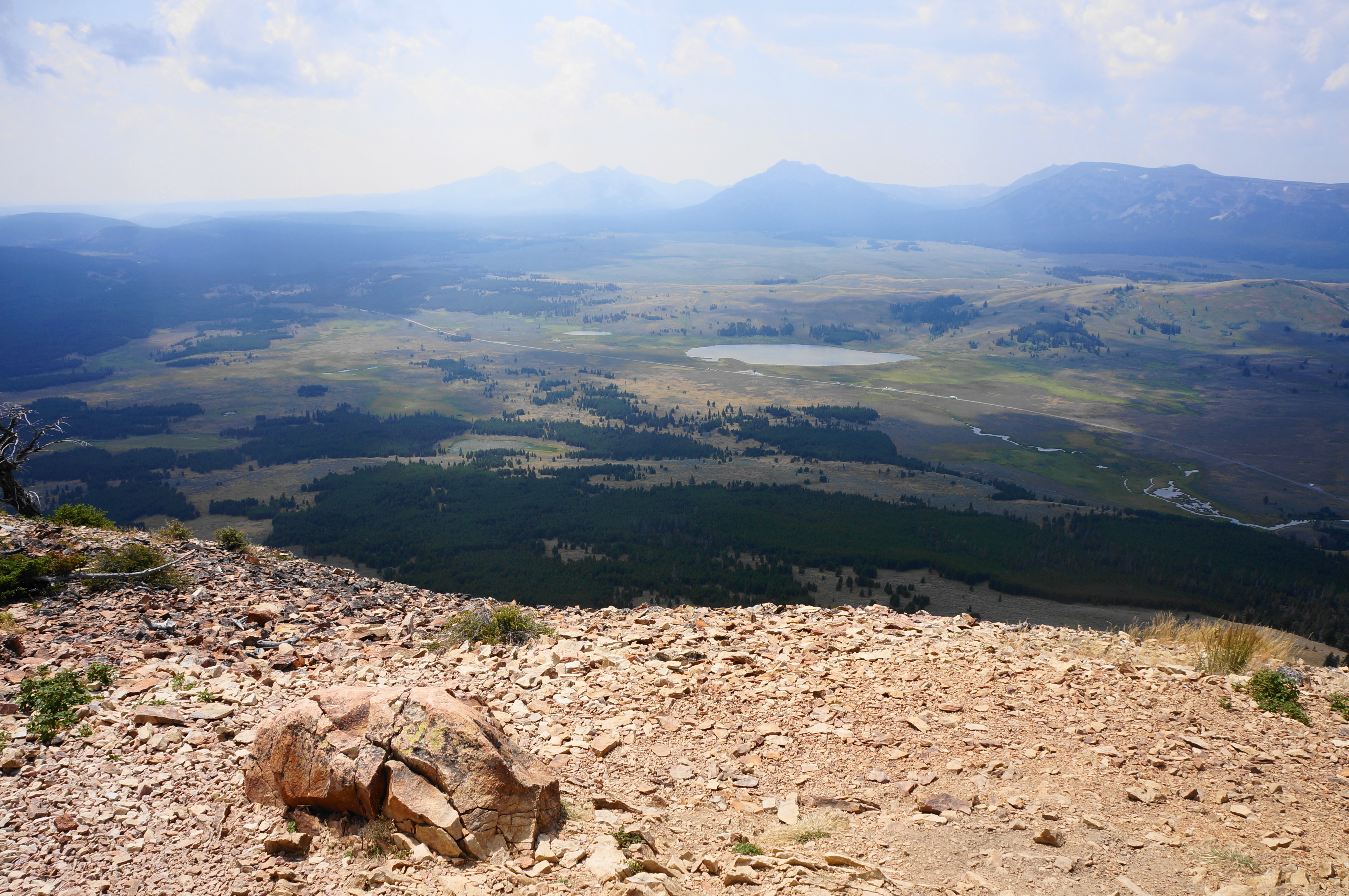 A real photograph from the Bunsen Peak trail in Yellowstone showing a rocky ridge with hikers looking out over a broad valley and distant mountains under a clear sky