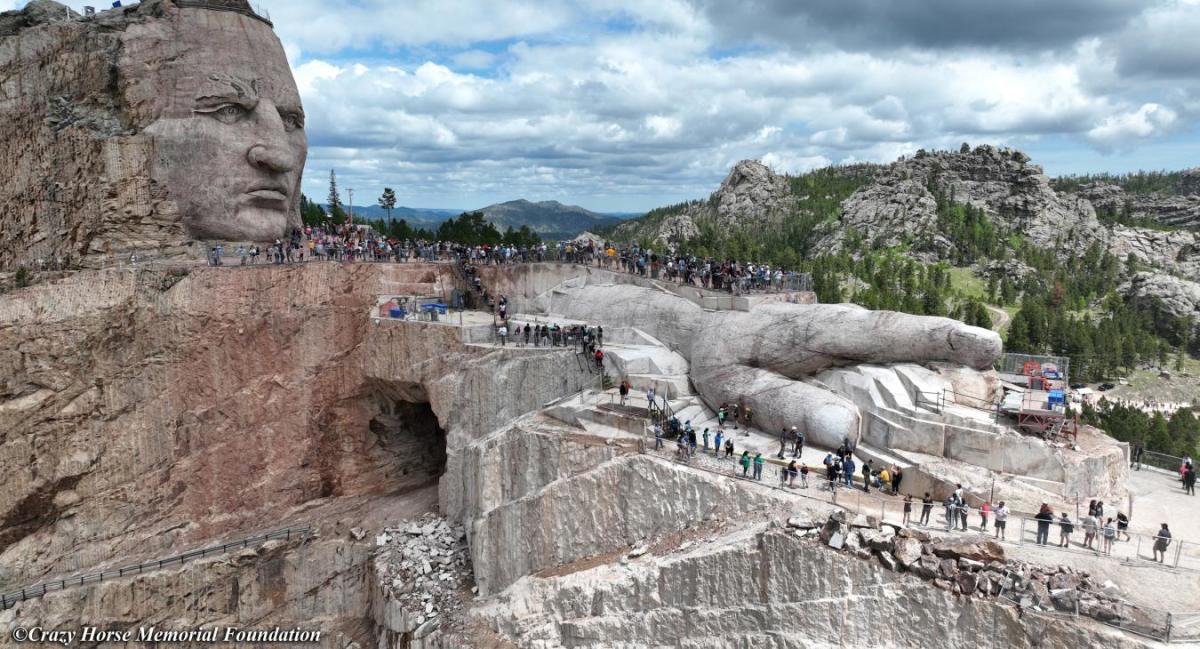 A real photograph from the Crazy Horse Memorial viewing terrace with the mountain carving site visible across the valley and visitors looking out