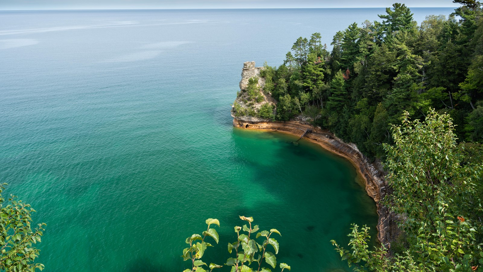 A real photograph from the Miners Castle overlook showing a dramatic sandstone cliff formation above Lake Superior with green forest on top and blue water below