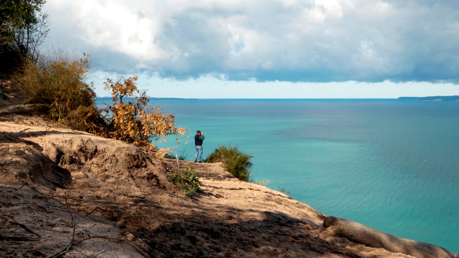 A real photograph from the Pyramid Point overlook showing steep forested slopes dropping to a bright turquoise Lake Michigan with a sandy shoreline below