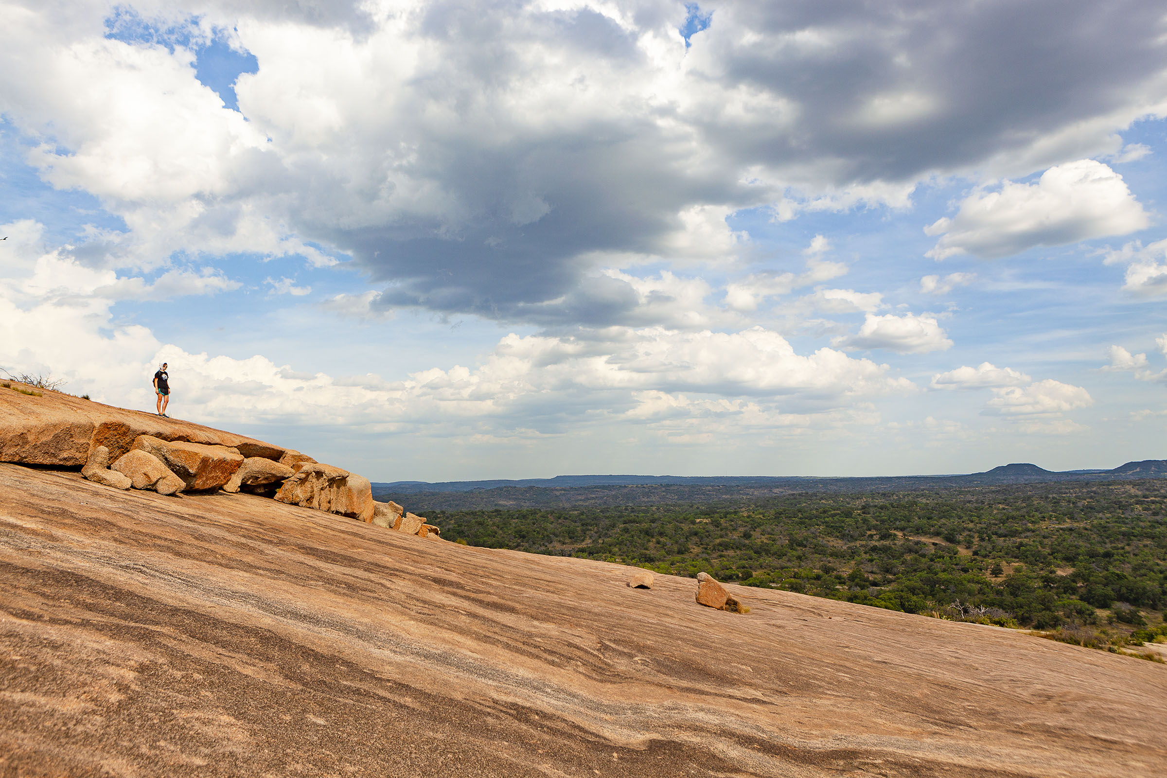 A real photograph from the top of Enchanted Rock in Texas looking out over rolling Hill Country, with pink granite in the foreground and distant green hills under a clear blue sky