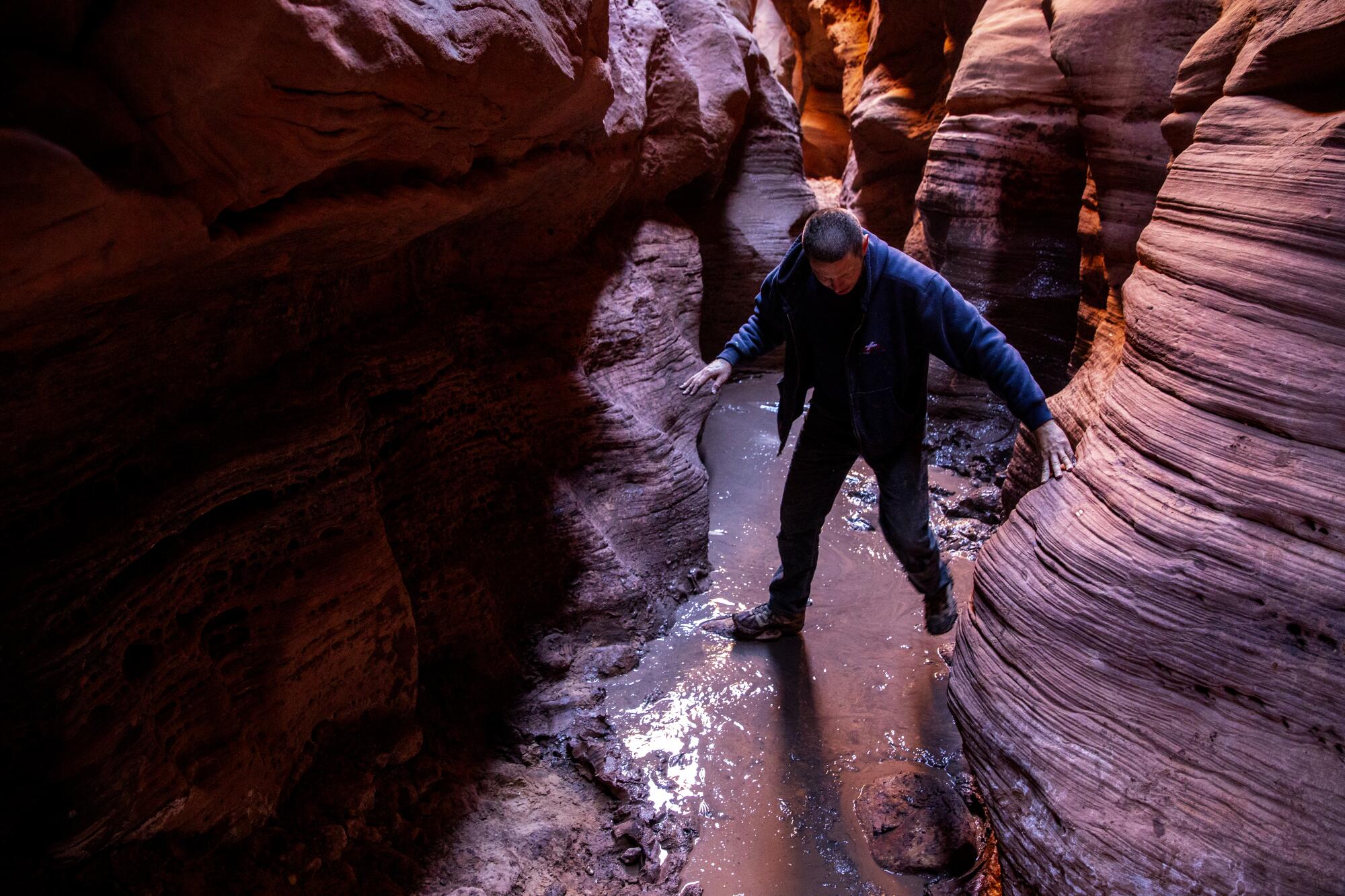 A real photograph inside Buckskin Gulch showing a narrow sandstone slot with a shallow standing water pool reflecting canyon walls, with wet sand and rippled mud along the edges