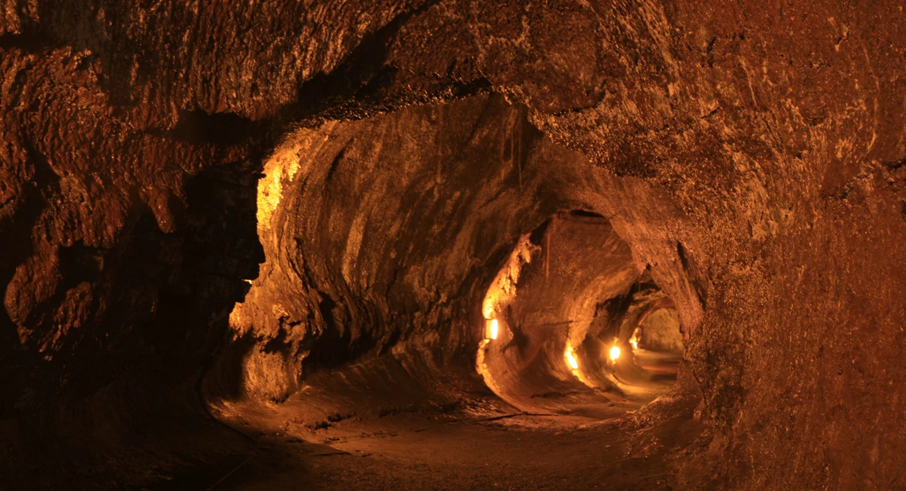 A real photograph inside Thurston Lava Tube in Hawaiʻi Volcanoes National Park, showing a dim tunnel with rough lava walls and a paved path lit by warm overhead lights
