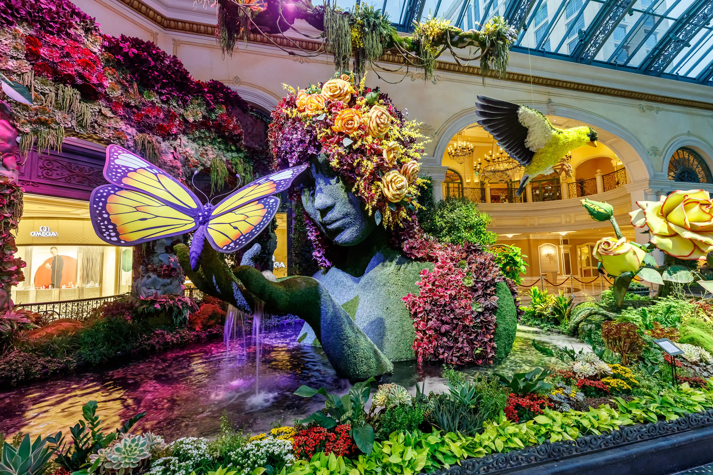 A real photograph inside the Bellagio Conservatory in Las Vegas with a large seasonal floral sculpture, lush greenery, and visitors walking along the path
