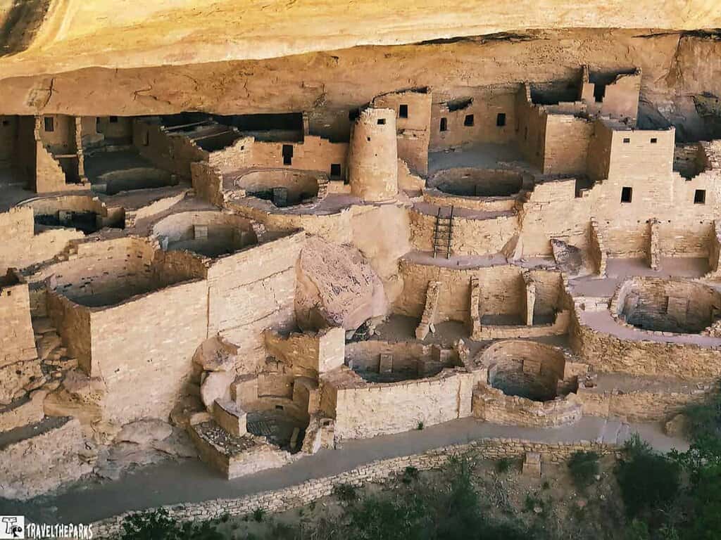 A real photograph looking across the sandstone alcove at Cliff Palace in Mesa Verde National Park, with stacked stone rooms and kivas in warm afternoon light