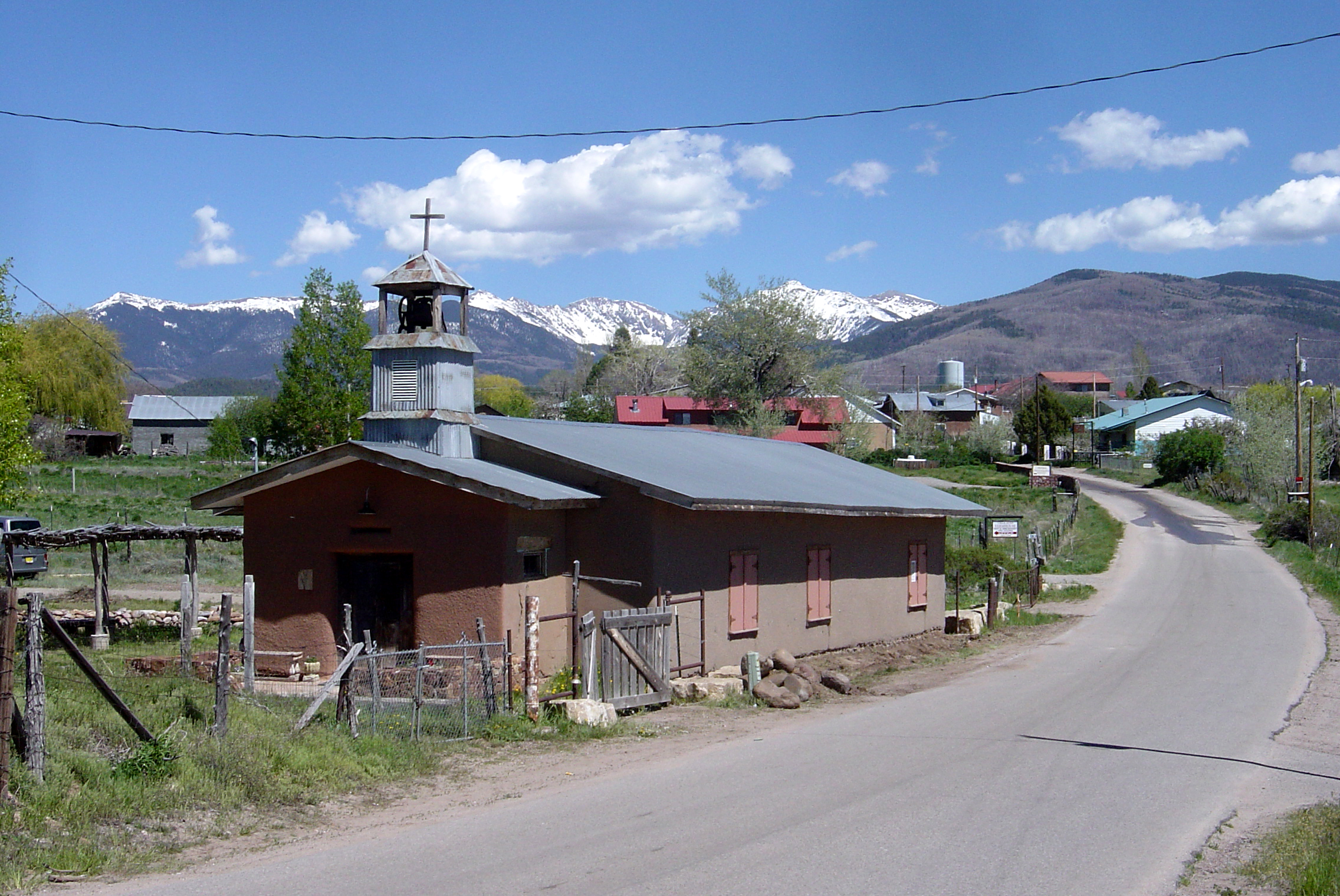 A real photograph looking down a narrow road near Truchas with adobe homes and a sweeping view of the Sangre de Cristo Mountains in the distance