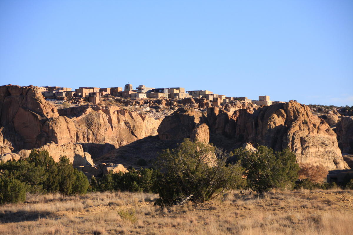 A real photograph of Acoma Pueblo Sky City on top of a sandstone mesa in New Mexico, with adobe buildings catching warm afternoon light and wide desert plains in the background