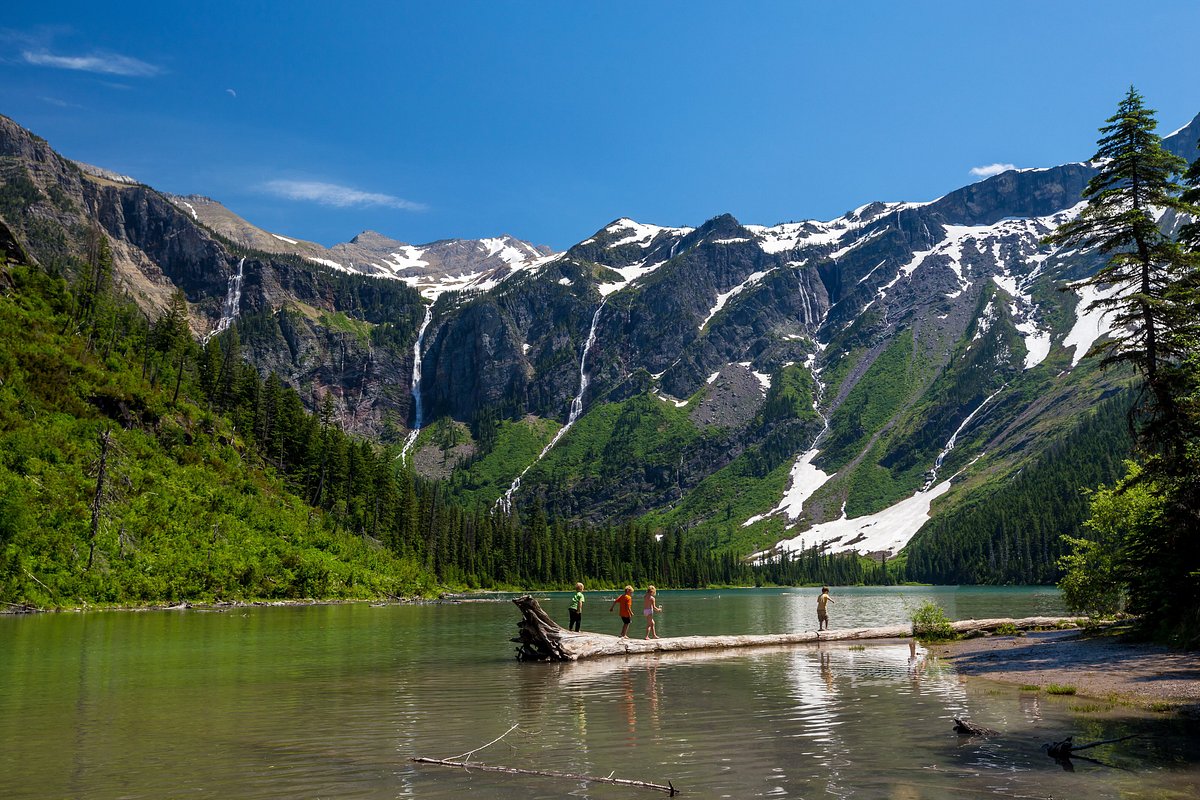 A real photograph of Avalanche Lake with a tall waterfall cascading down a mountainside into a clear lake, hikers resting on the rocky shoreline, evergreen forest around