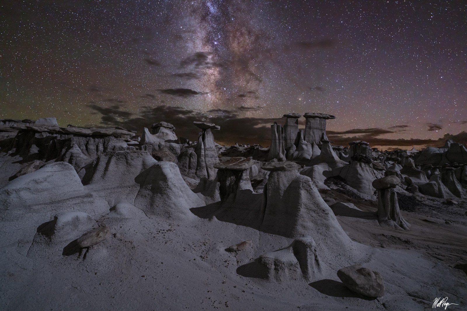 A real photograph of Badlands rock formations silhouetted under a clear night sky filled with stars, with a faint glow on the horizon