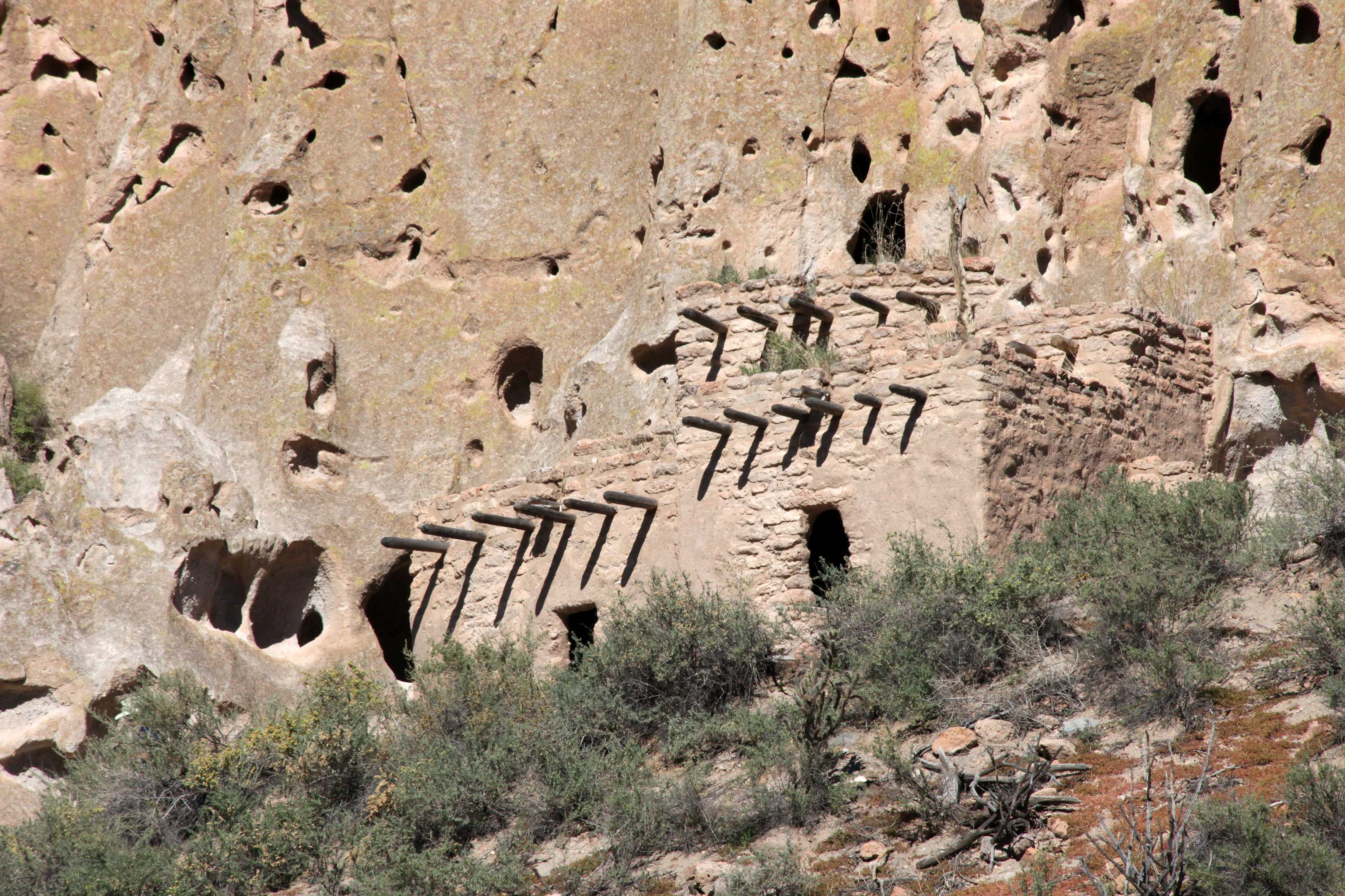 A real photograph of Bandelier National Monument in New Mexico showing tuff rock canyon walls with ancestral cliff dwelling openings and a trail below