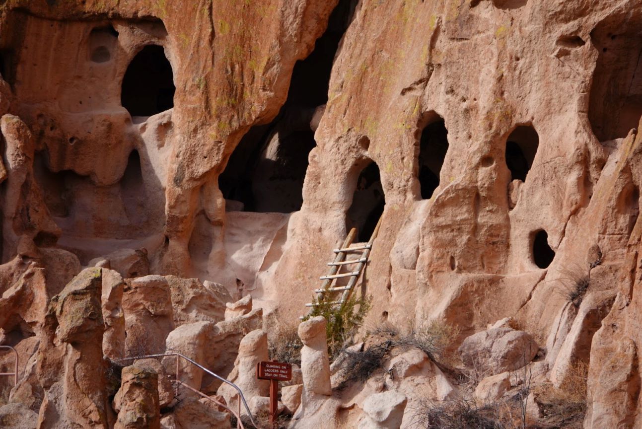 A real photograph of Bandelier National Monument showing a ladder leading up to a carved alcove in tan volcanic tuff with stone masonry ruins below