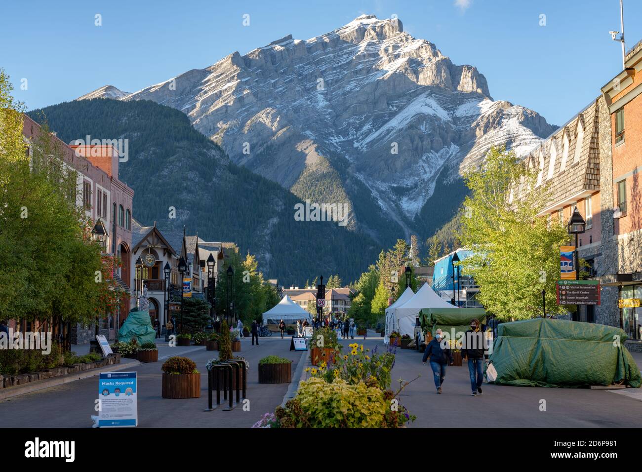 A real photograph of Banff Avenue on a clear summer morning, with Cascade Mountain rising at the end of the street and people walking past shops and cafes