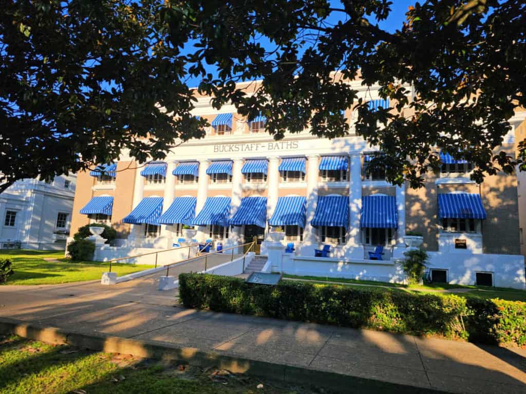 A real photograph of Bathhouse Row along Central Avenue in Hot Springs, Arkansas, with the historic brick and stone bathhouse buildings lined up beside the sidewalk on a bright daytime scene