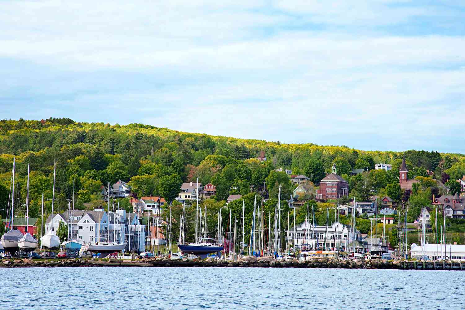 A real photograph of Bayfield, Wisconsin with sailboats and tour boats moored in the harbor, historic storefronts on the shoreline, and a calm Lake Superior backdrop in late-summer light