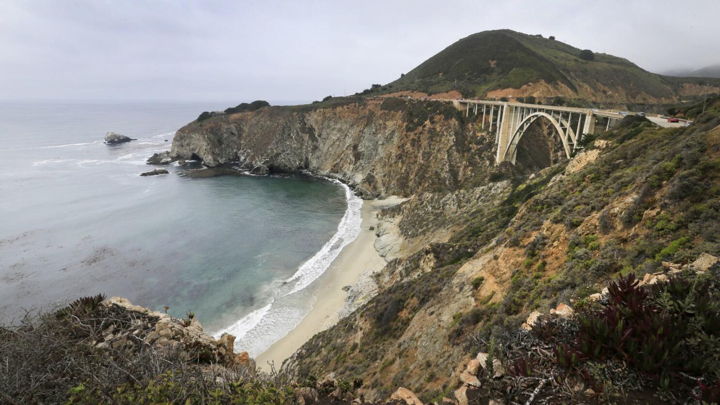 A real photograph of Bixby Creek Bridge in Big Sur on a clear morning, with the arched concrete bridge spanning a rocky canyon and the Pacific Ocean stretching behind it