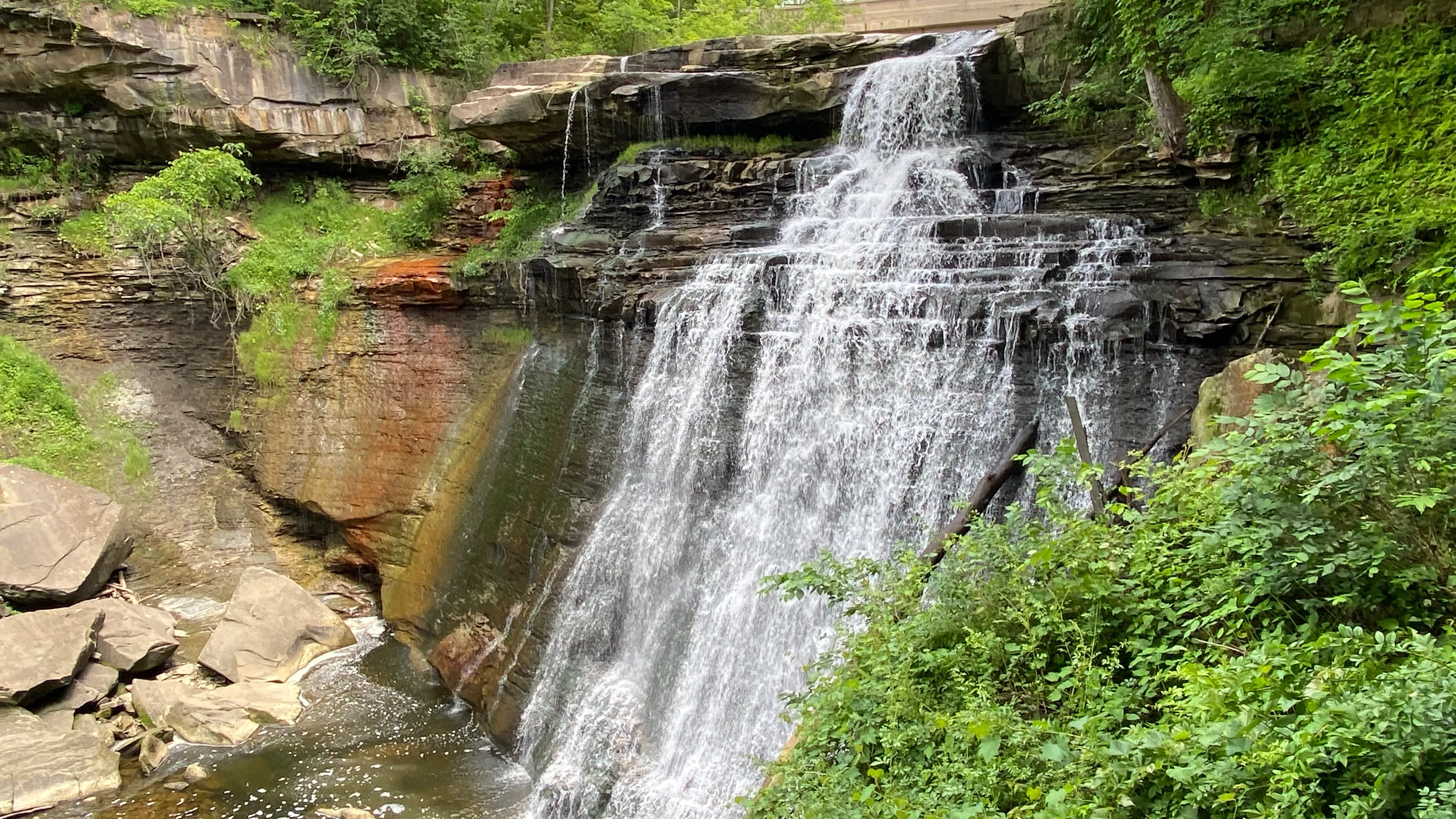 A real photograph of Brandywine Falls dropping into a rocky gorge surrounded by green trees in Cuyahoga Valley National Park on a bright spring morning
