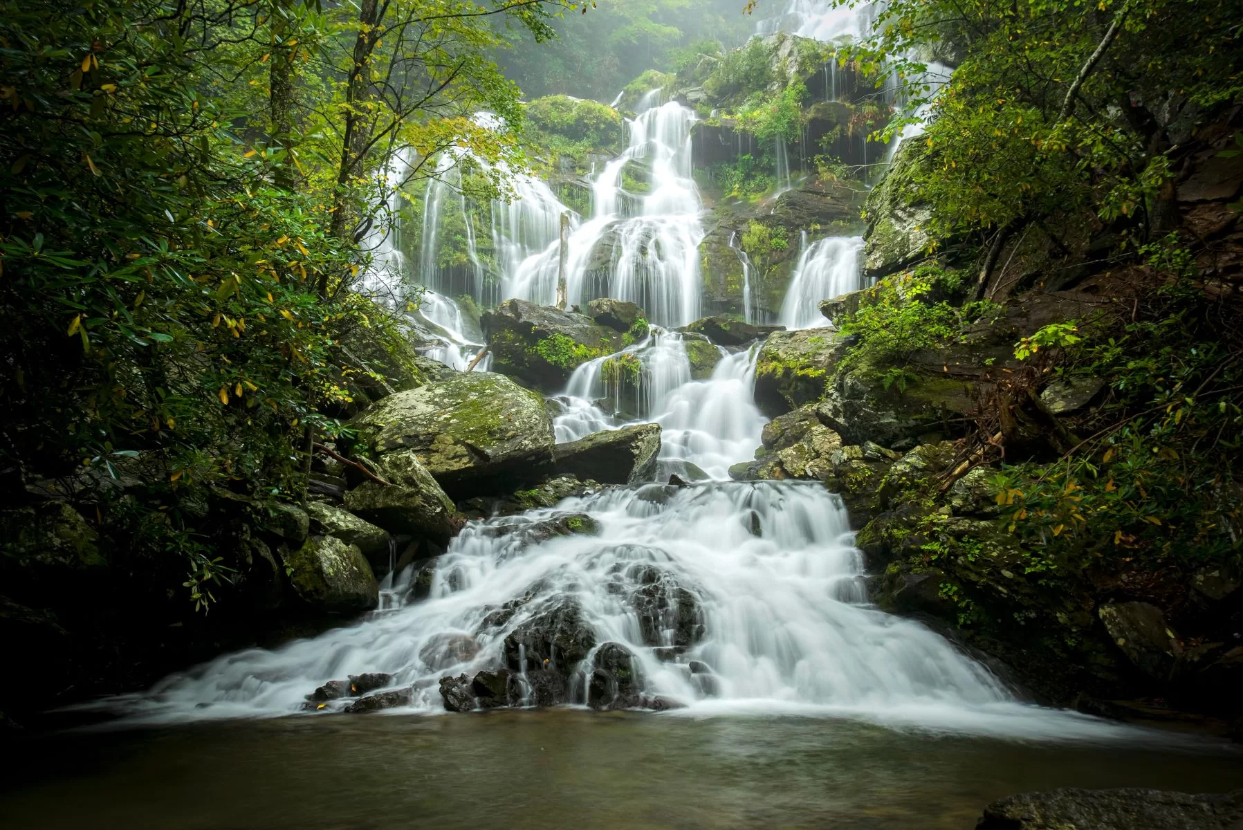 A real photograph of Catawba Falls near Asheville with water cascading over rock ledges into a clear pool, lush green forest framing the scene, long-exposure waterfall photography style