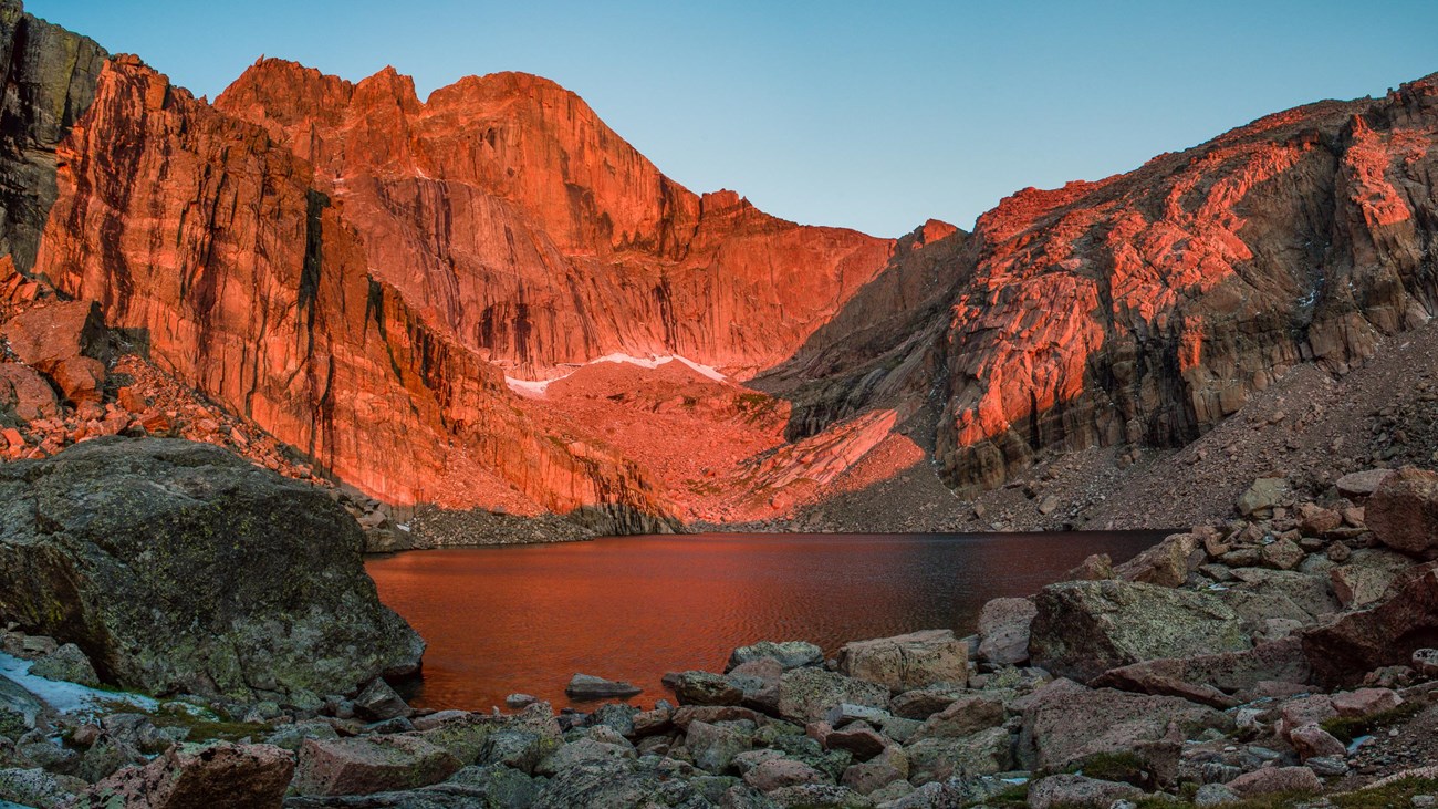 A real photograph of Chasm Lake with dark blue water in a rocky basin and the steep east face of Longs Peak rising sharply behind it