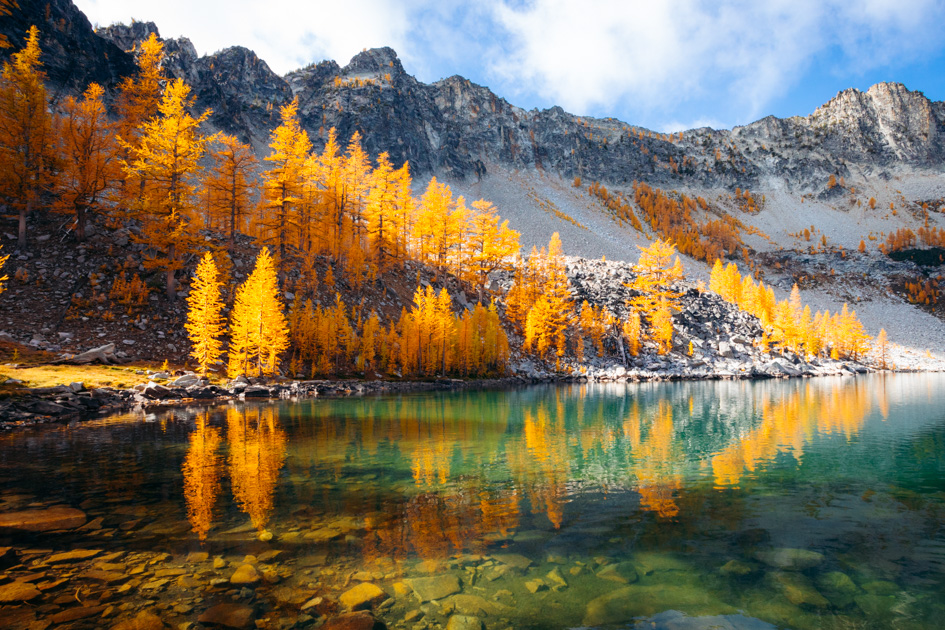 A real photograph of Colchuck Lake in Washington in early autumn, with golden larch trees near the shore, turquoise water, and jagged granite peaks rising behind the lake