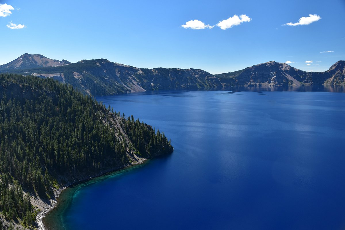 A real photograph of Crater Lake viewed from a Rim Drive overlook in summer, with deep blue water, Wizard Island centered in the lake, and a stone guardrail in the foreground