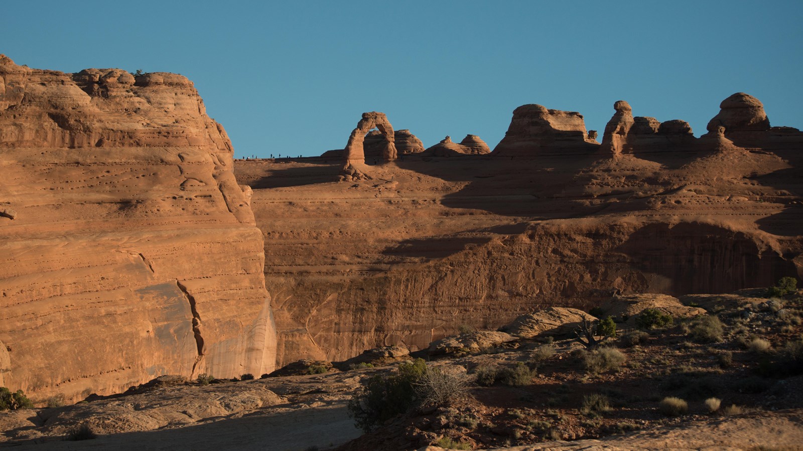 A real photograph of Delicate Arch framed against a blue sky with red sandstone and a few hikers visible on the slickrock trail in Arches National Park