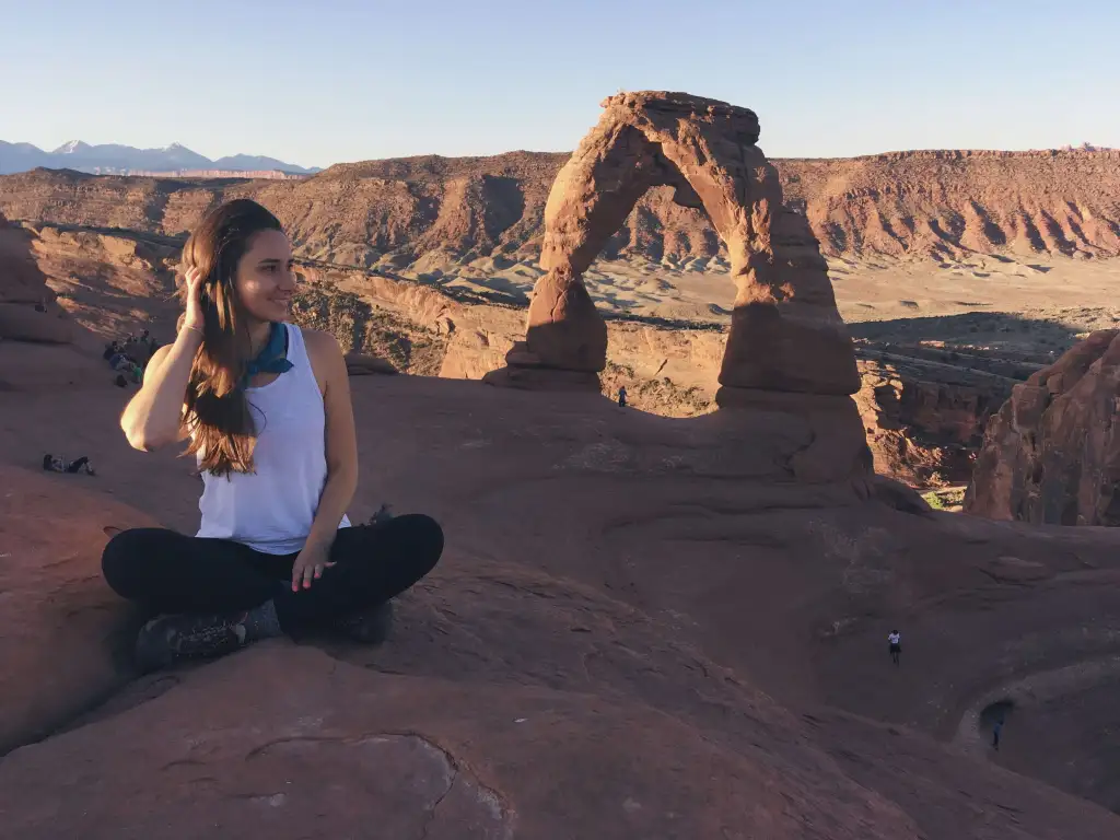 A real photograph of Delicate Arch in Arches National Park glowing orange at sunrise, with a few hikers standing on the slickrock viewpoint in the distance