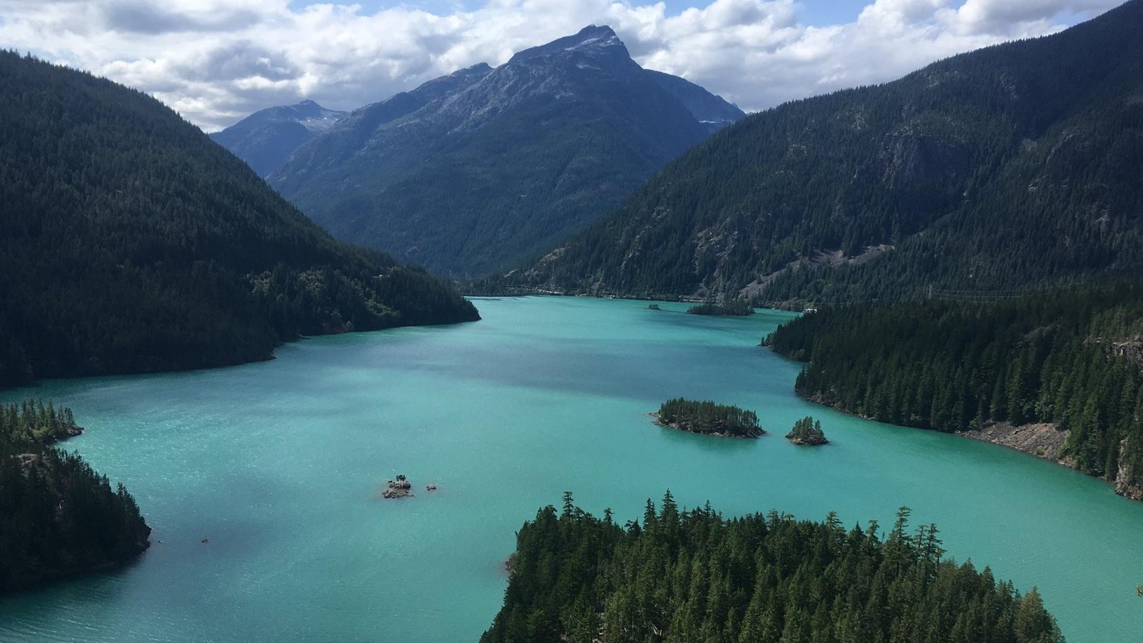A real photograph of Diablo Lake seen from a high roadside overlook, with vivid turquoise water, dark evergreen forests, and jagged North Cascades peaks under a clear summer sky