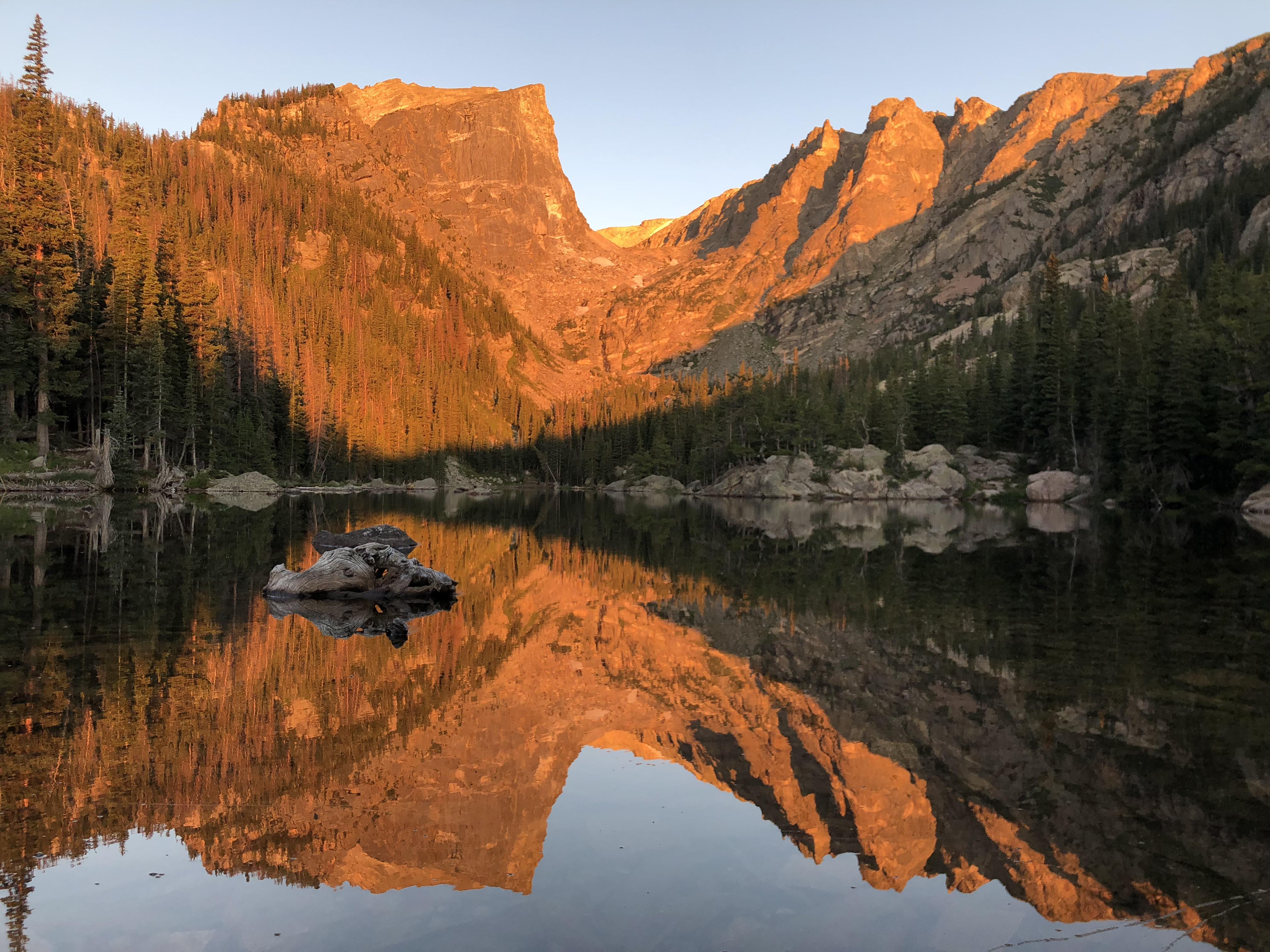 A real photograph of Dream Lake in Rocky Mountain National Park at sunrise, with still water reflecting a rocky peak and evergreen shoreline