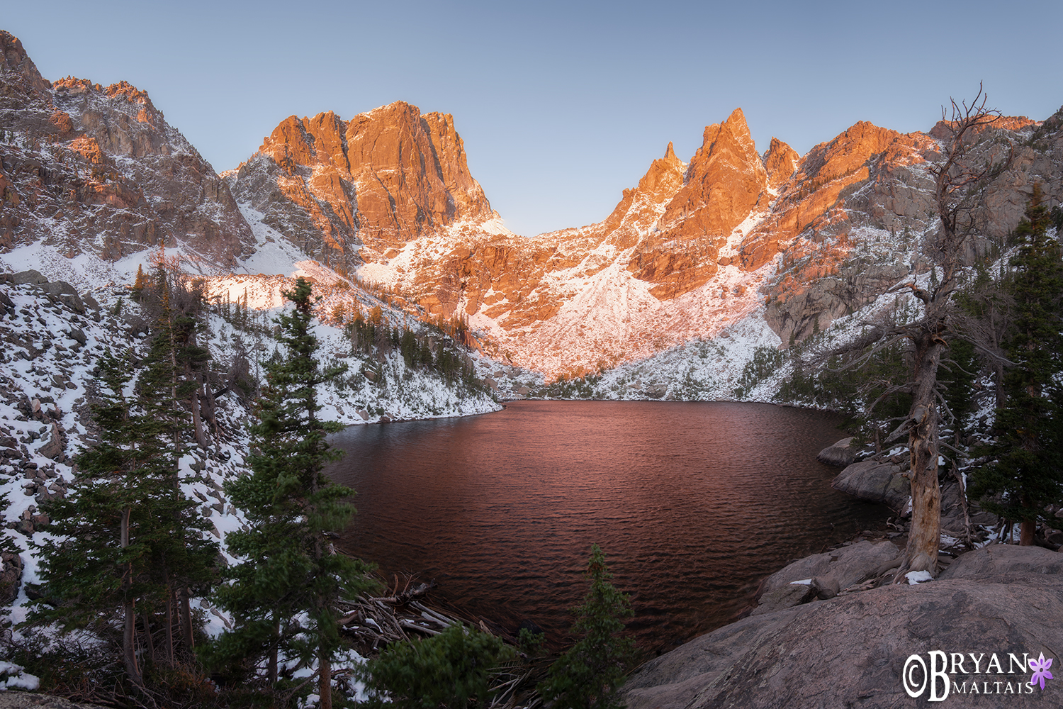 A real photograph of Emerald Lake in Rocky Mountain National Park at sunrise, with calm water reflecting Hallett Peak and Flattop Mountain, and a rocky shoreline in the foreground