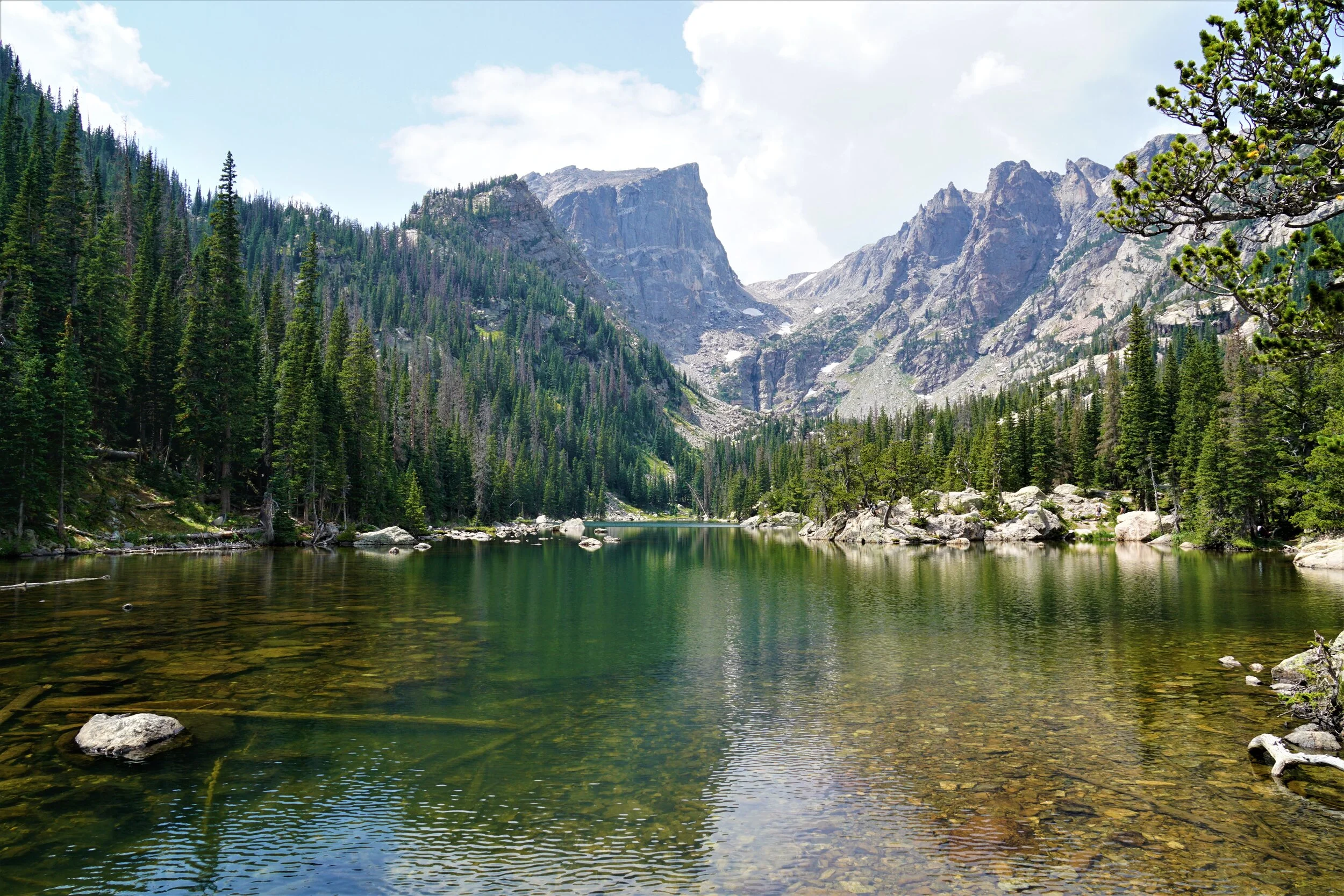 A real photograph of Emerald Lake in Rocky Mountain National Park on a clear summer day, with hikers resting on rocks near the shoreline and rugged mountain peaks rising behind the water
